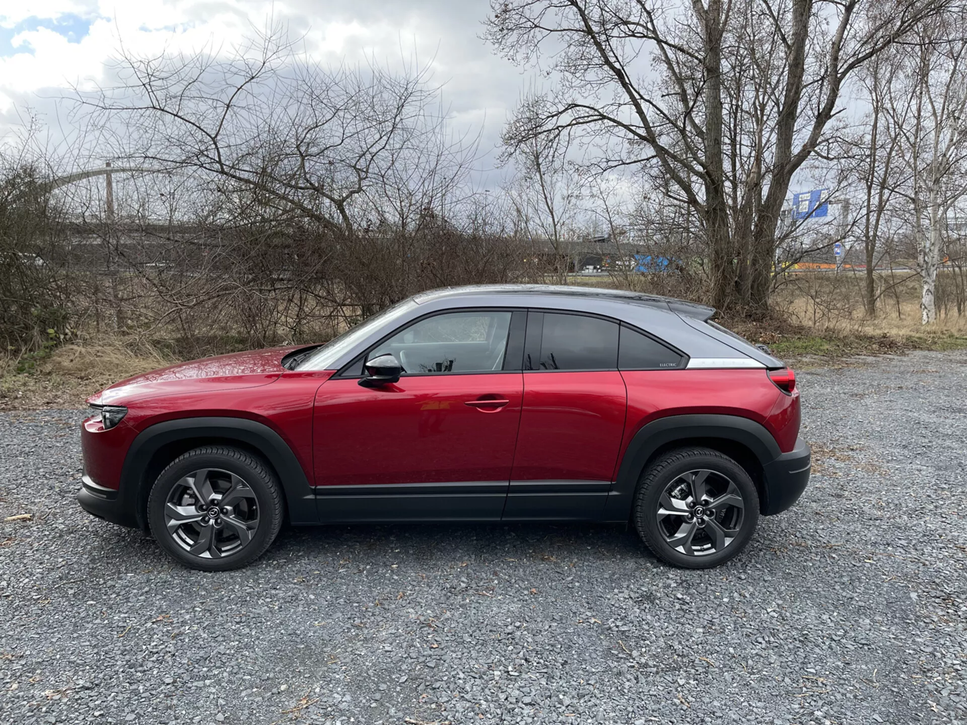 Perfil lateral do SUV crossover Mazda MX-30 2020 vermelho em cascalho com vegetação escassa e céu nublado, apresentando linha de cintura alta, linha de teto inclinada estilo cupê, linha de caráter dinâmico, rodas de liga leve com múltiplos raios com revestimento plástico preto, e possíveis características tecnológicas avançadas como sistemas ADAS, iluminação LED, entrada sem chave, sistema de monitoramento da pressão dos pneus TPMS e sistema de infotainment.