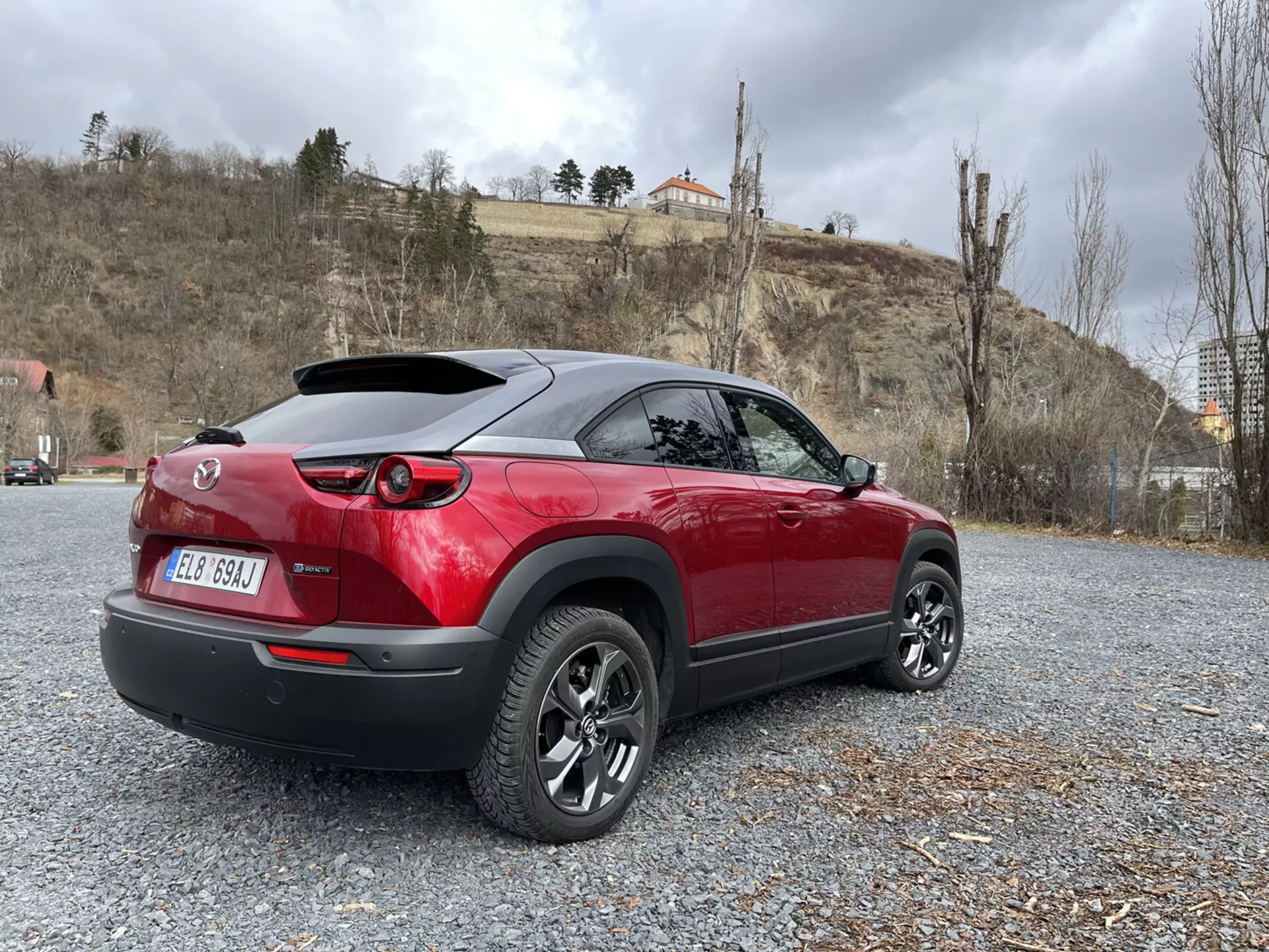 Crossover moderno Mazda MX-30 2020 estacionado em cascalho, exibindo seu design esportivo em dois tons de vermelho e preto, luzes traseiras LED, spoiler traseiro e rodas de liga leve multirradiadas contra um fundo de natureza e arquitetura