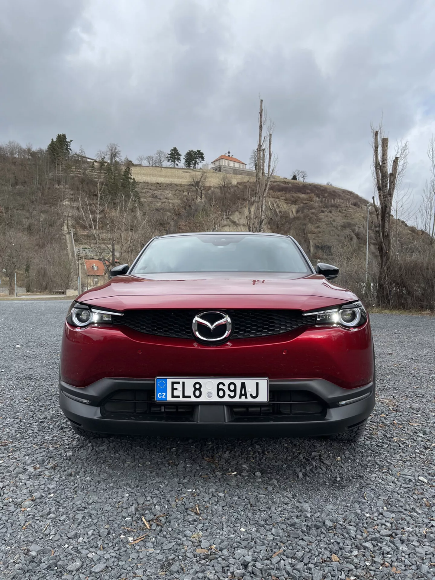 Vista frontal de um Mazda MX-30 de 2020 vermelho, destacando a grelha, o pára-choques e os faróis LED do carro sobre uma superfície de cascalho contra um fundo de céu nublado com vegetação e um edifício