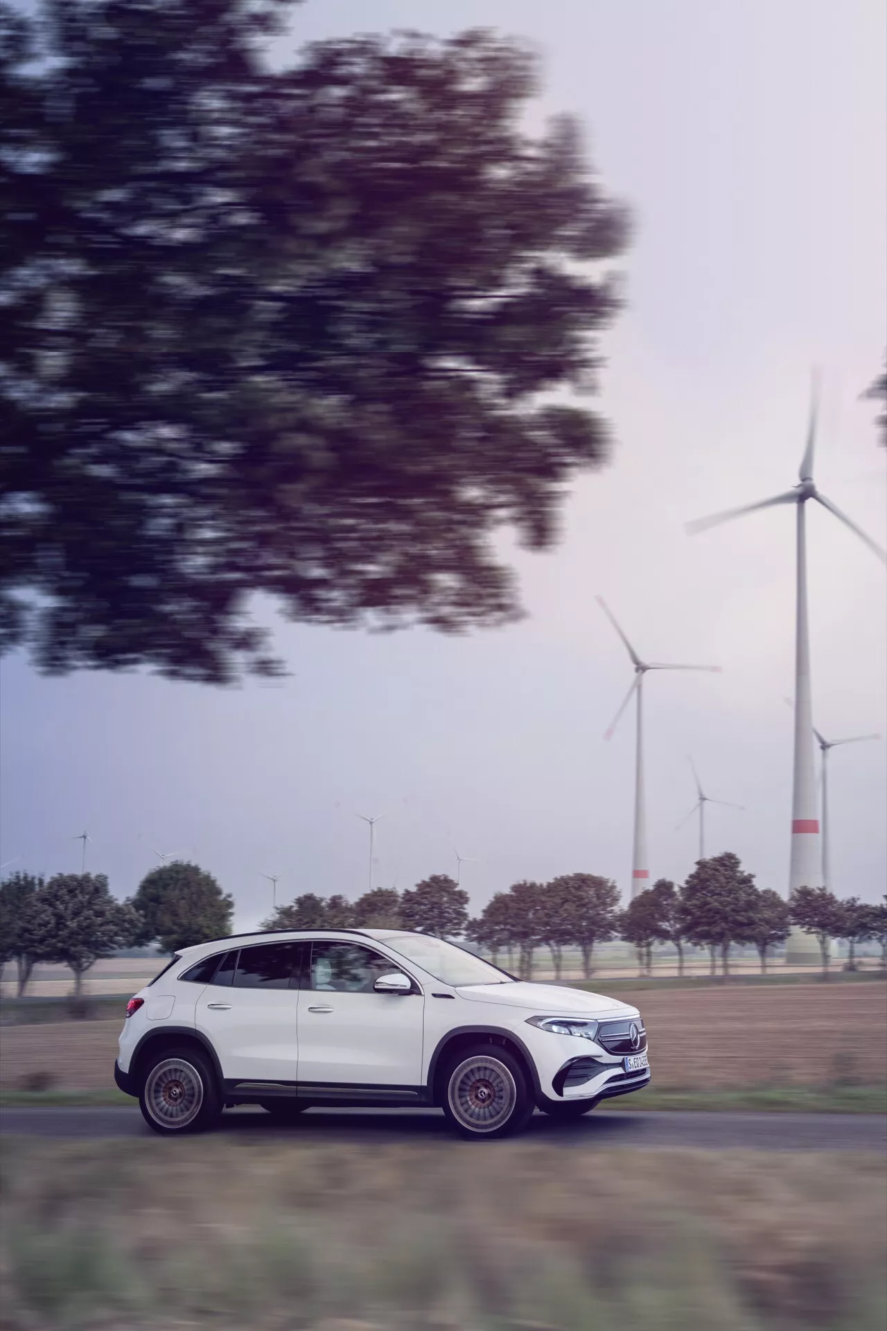 Mercedes-Benz EQA 2021 em movimento, vista de perfil lateral exibindo design de luxo e aerodinâmica, com turbinas eólicas ao fundo indicando tecnologia ecologicamente correta