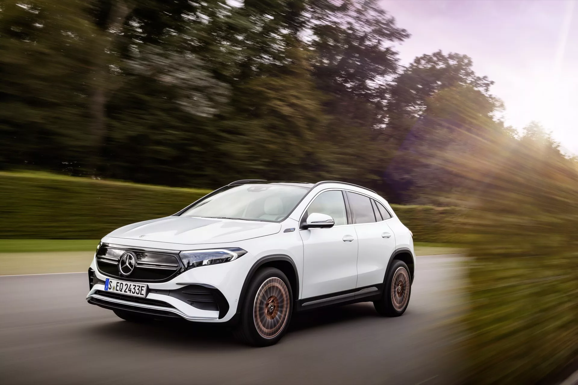 SUV branco Mercedes-Benz EQA 2021 em movimento, vista de três quartos frontal destacando a postura dinâmica, design aerodinâmico, faróis LED e rodas de liga leve distintas em uma estrada cênica