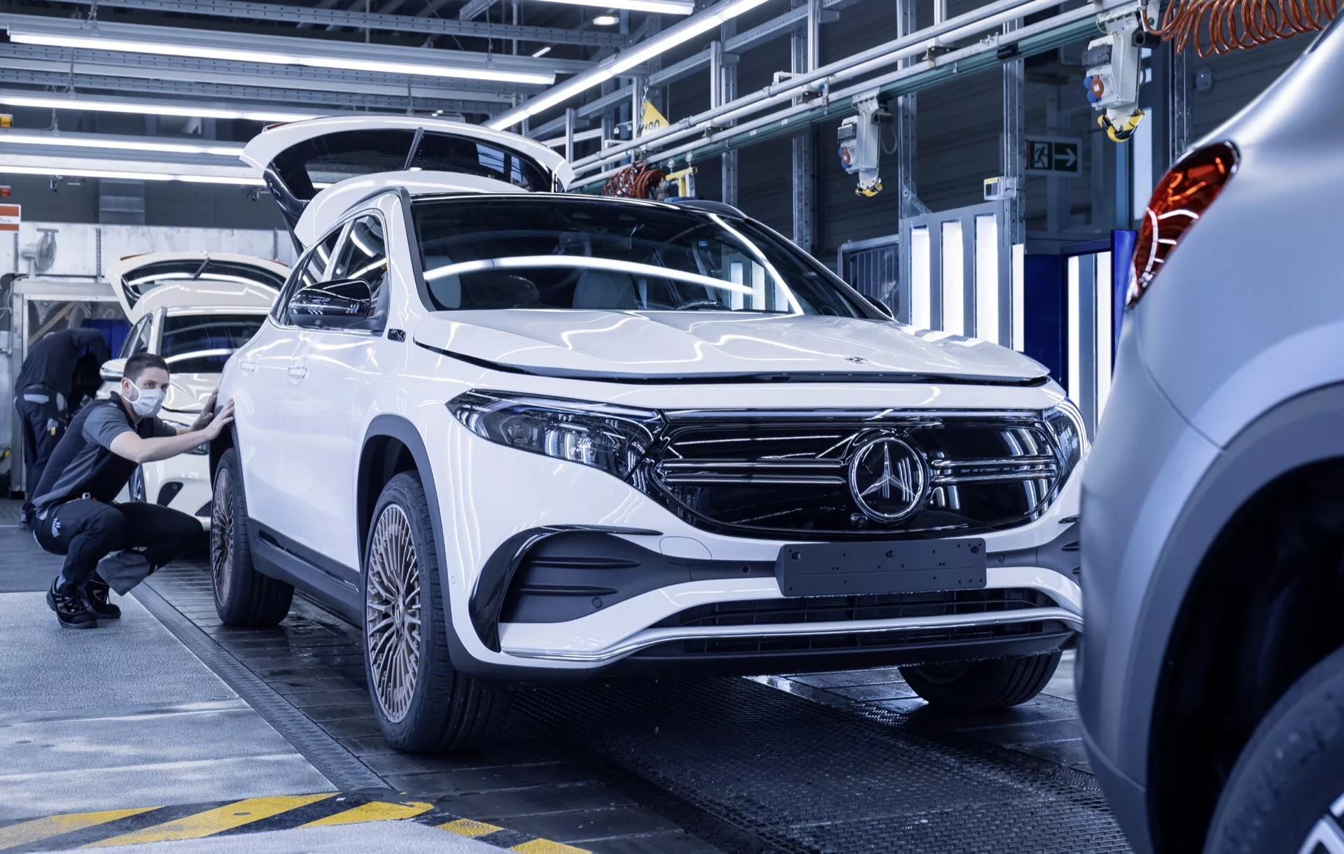Mercedes-Benz EQA 2021 em branco, exibindo um design moderno de SUV com uma vista angular frontal e lateral dentro de uma instalação de montagem ou inspeção de automóveis, destacando os faróis LED elegantes, para-choque dianteiro aerodinâmico e emblema proeminente na grade.