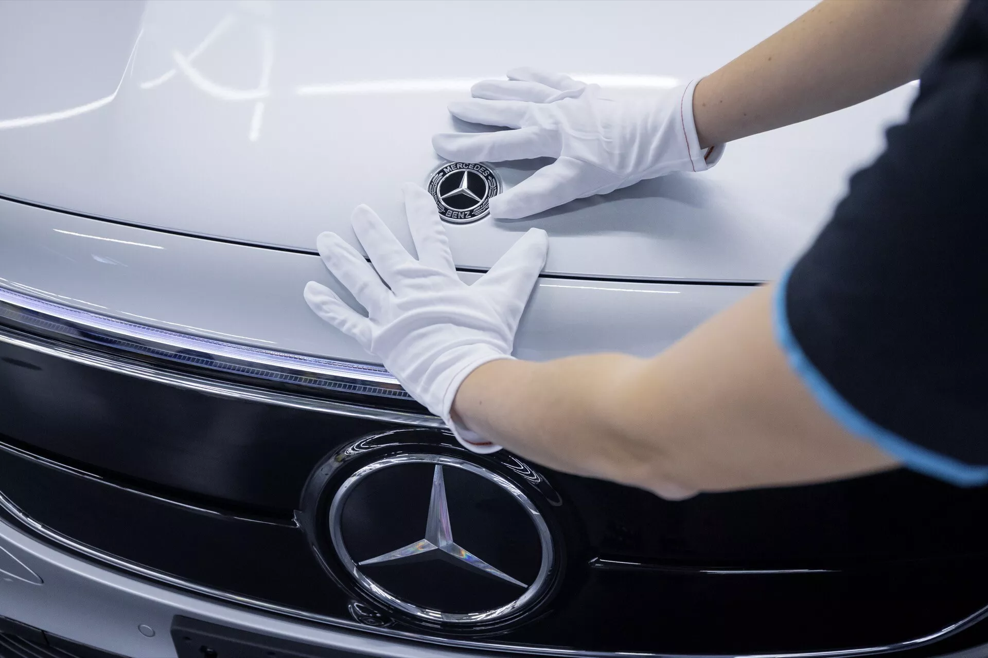 Vista de perto da grade e emblema do Mercedes-Benz EQA 2021 com mãos em luvas brancas tocando, destacando detalhes em cromo e tecnologia de faróis LED.