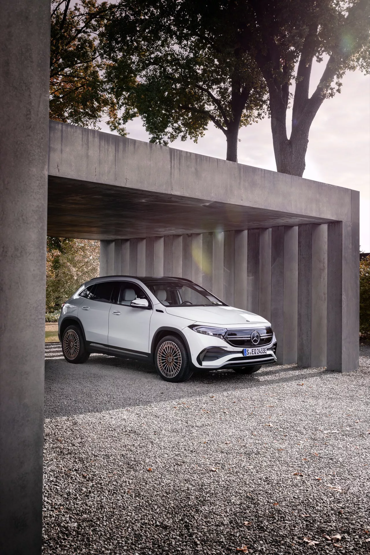 Mercedes-Benz EQA 2021 estacionado sob uma estrutura de concreto minimalista, exibindo seus faróis LED elegantes, grade frontal distintiva com emblema, carroceria branca aerodinâmica e rodas de liga intrincadas de uma vista frontal de três quartos em ângulo baixo.