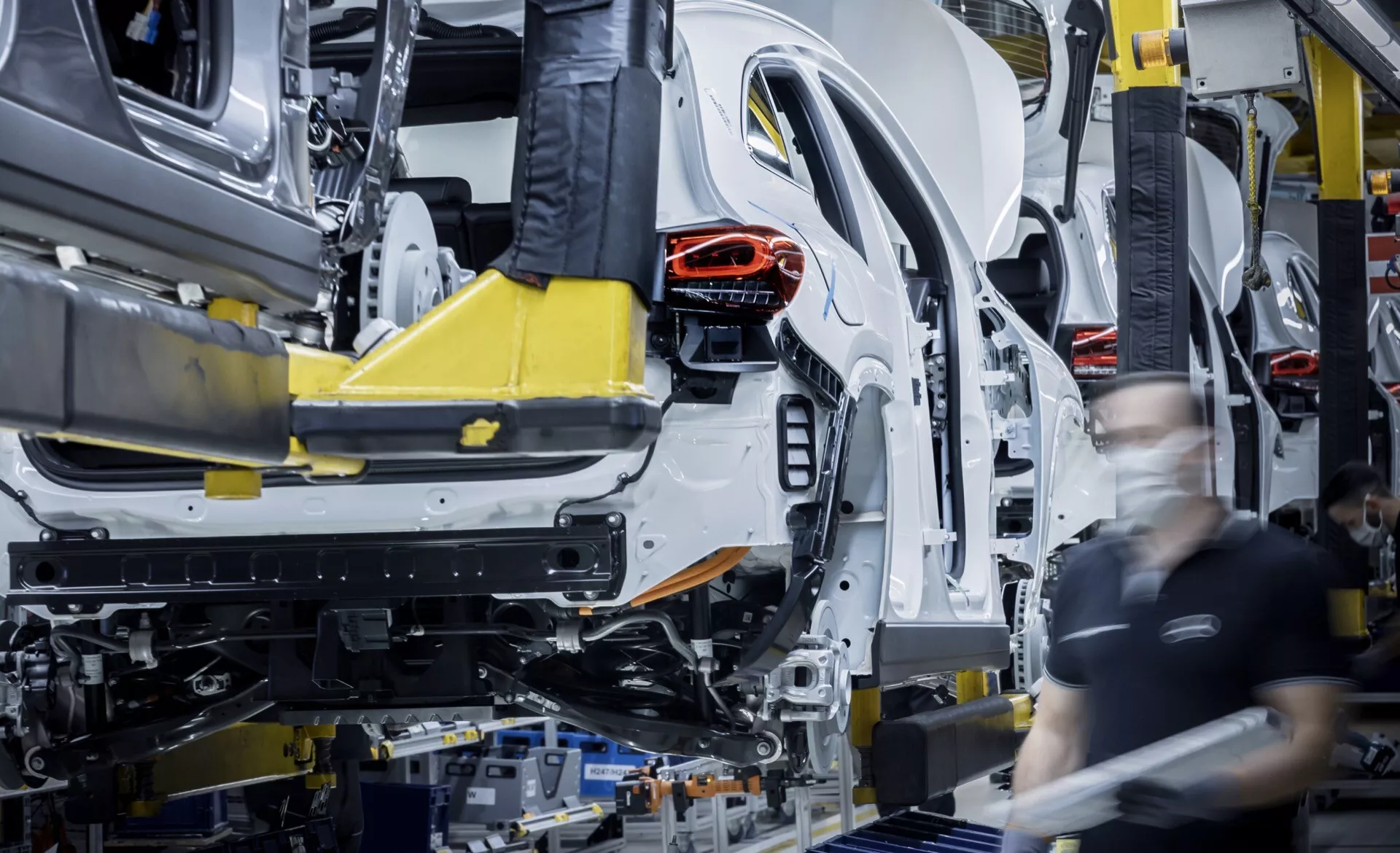 Montagem em estágio inicial do Mercedes-Benz EQA 2021 na linha de fábrica, mostrando a carroceria e chassi unibody suportados por braços robóticos automatizados com estrutura traseira visível e trabalhadores de montagem em segundo plano