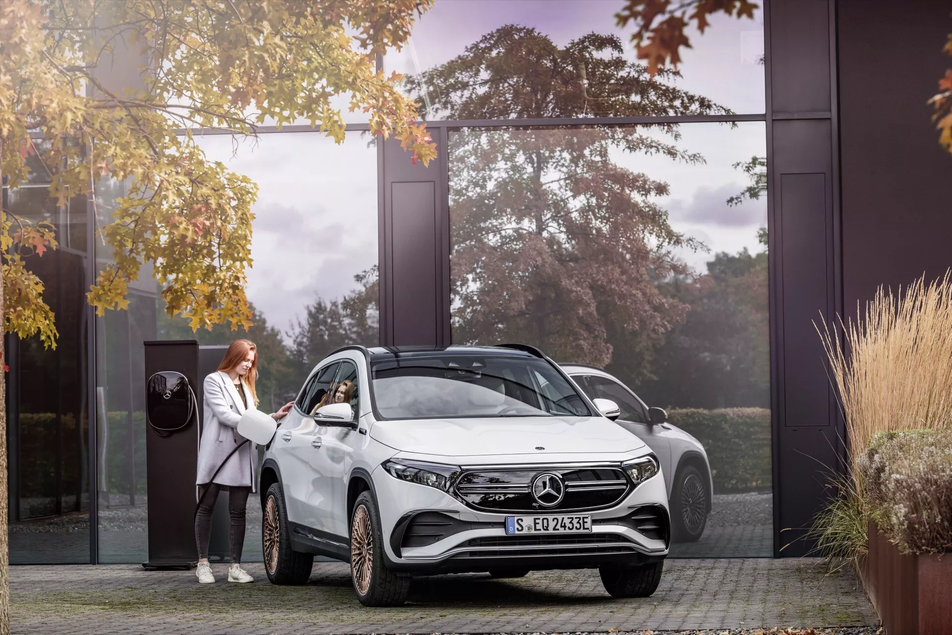 SUV Mercedes-Benz EQA 2021 em entrada de residência, com faróis LED, rodas de liga leve, emblema elétrico e pessoa demonstrando entrada sem chave, rodeado por árvores de outono