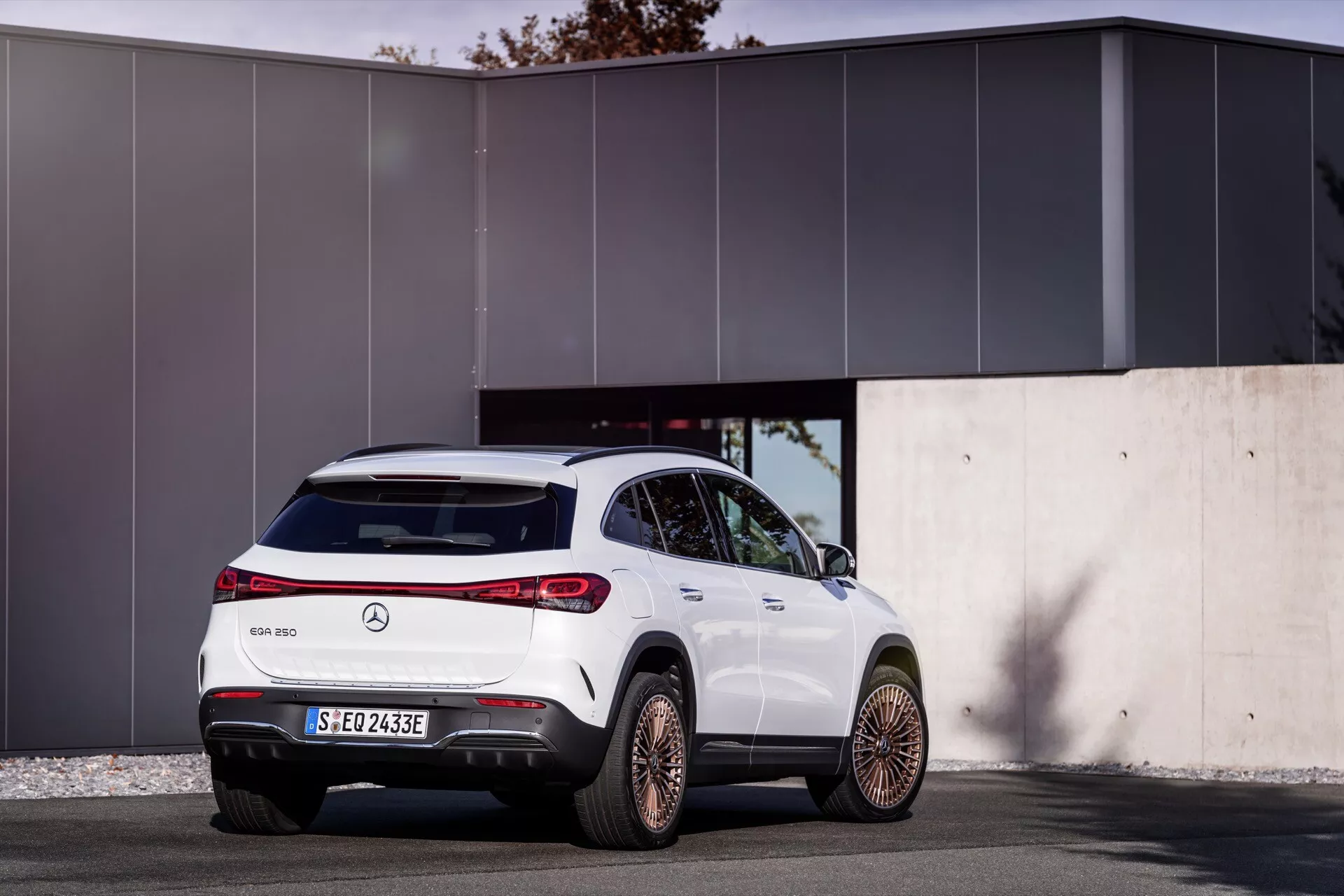 Vista traseira de três quartos de um SUV Mercedes-Benz EQA 2021 branco estacionado ao ar livre apresentando design moderno com linhas contornadas, luzes traseiras distintas envolventes, para-choque traseiro esportivo com difusor, rodas de liga leve com cinco raios divididos, emblema de veículo elétrico, sensores de estacionamento traseiro, luz de stop elevada, acabamento brilhante com detalhes em cromo, contra um fundo de edifício moderno minimalista ao sol.