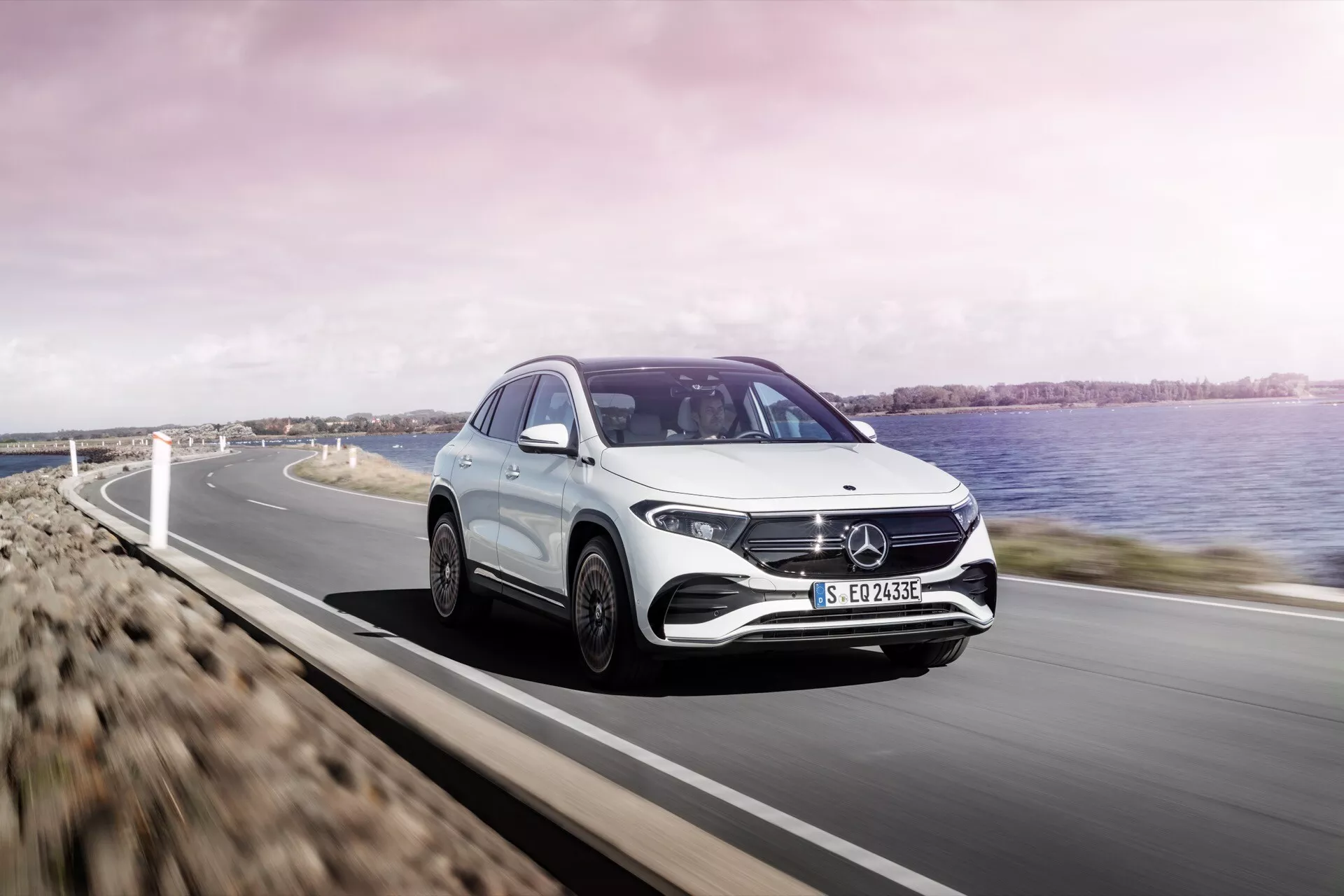 Mercedes-Benz EQA 2021 em movimento em uma estrada costeira, exibindo design aerodinâmico, faróis LED angulares e grade distintiva com emblema, visível contra um cenário pitoresco com água