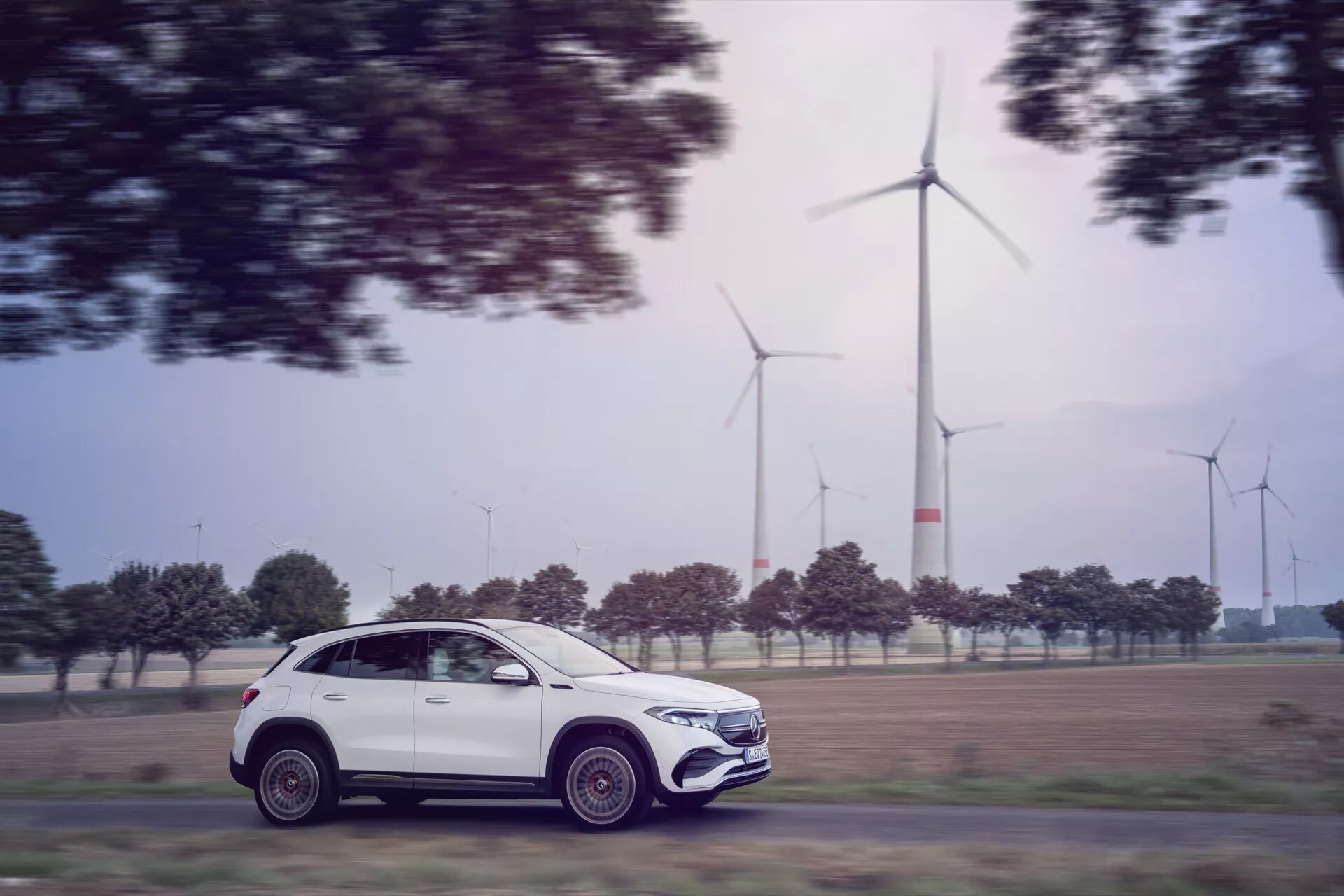 Mercedes-Benz EQA 2021 (243) em movimento capturado com técnica de fotografia panorâmica, exibindo seu perfil moderno de SUV, rodas de liga intrincadas e design ecológico contra um fundo de turbinas eólicas