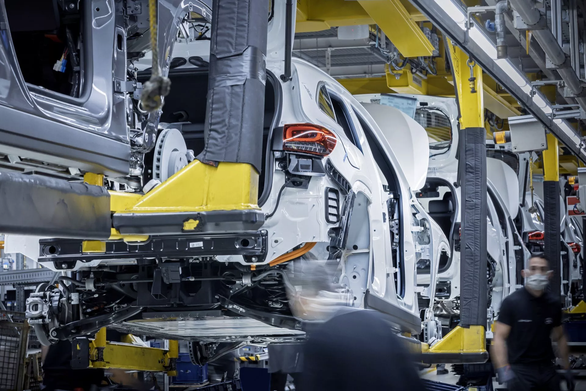 Montagem do perfil lateral do Mercedes-Benz EQA 2021 em andamento dentro da fábrica, exibindo metade traseira suspensa por braço robótico amarelo, estrutura unibody visível e componentes internos parciais.