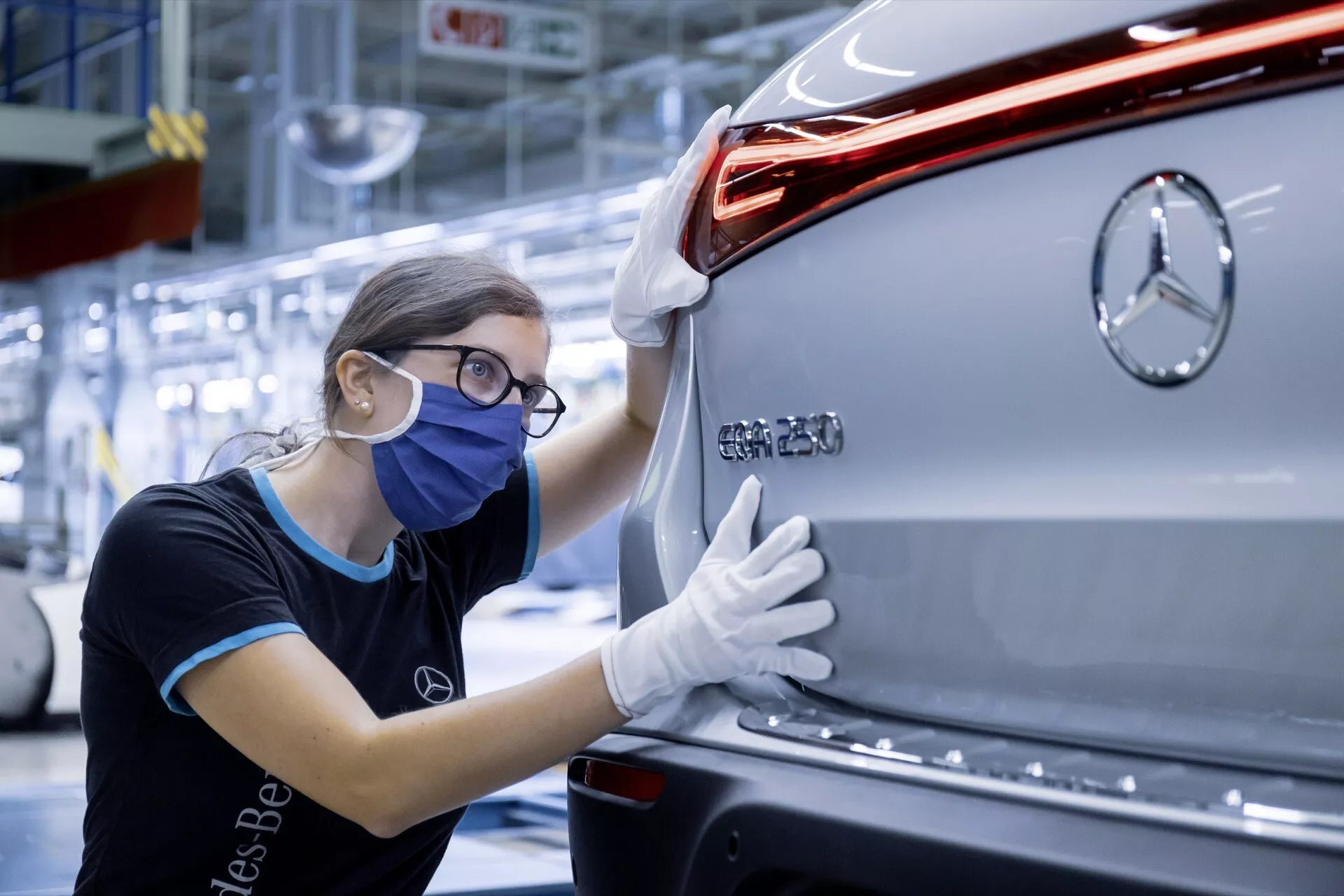 Vista traseira de um Mercedes-Benz EQC 2021 prata mostrando o emblema da marca, luz traseira LED moderna, design elegante do carro, e parachoque traseiro, com um trabalhador de controle de qualidade inspecionando o veículo em um ambiente de fábrica