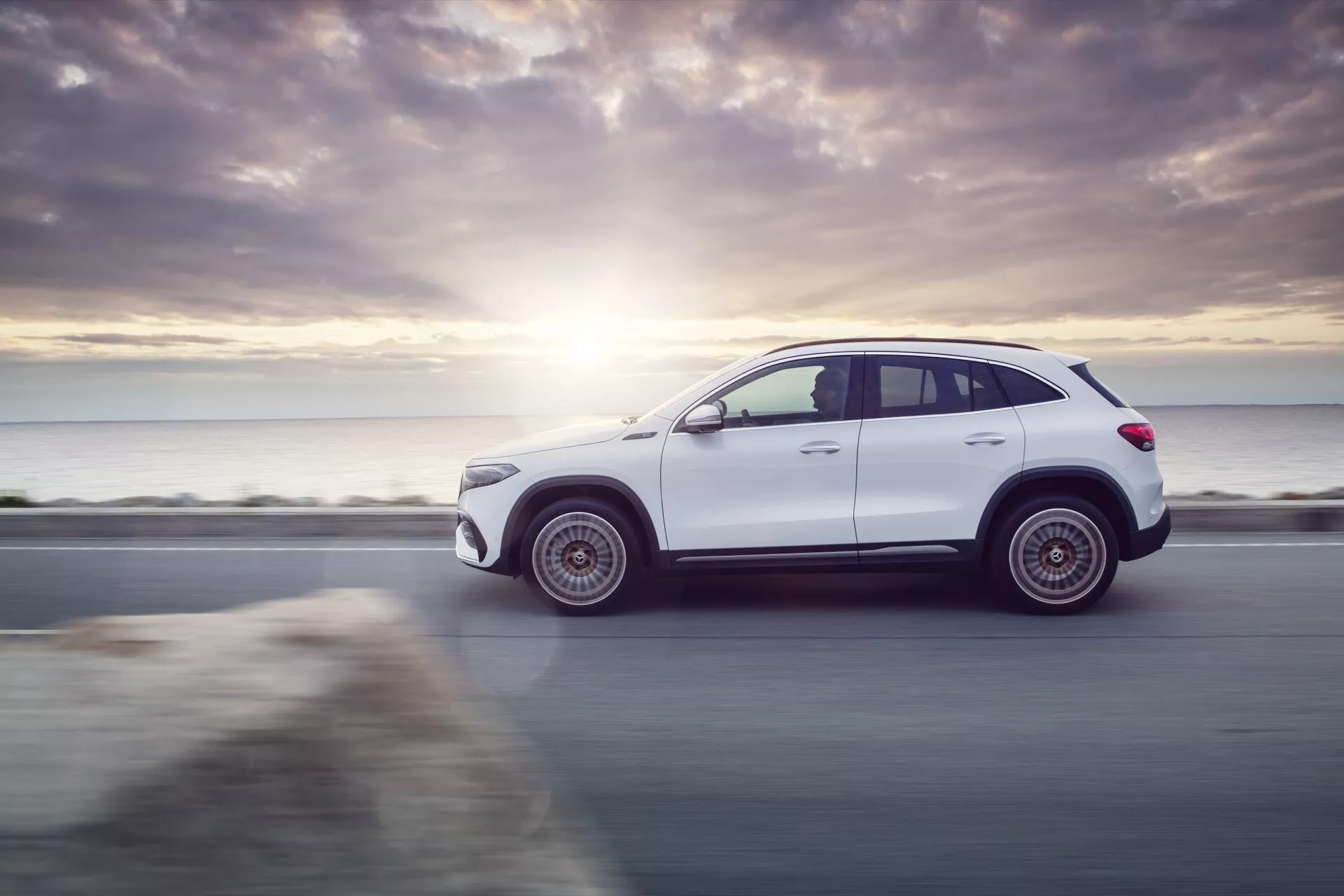 Perfil lateral dinâmico do SUV Mercedes-Benz EQA 2021 em branco, exibindo seu design elegante e postura robusta em uma estrada à beira-mar, destacado pelo efeito de flare do pôr do sol e características tecnológicas como rodas de liga leve e possível iluminação LED.