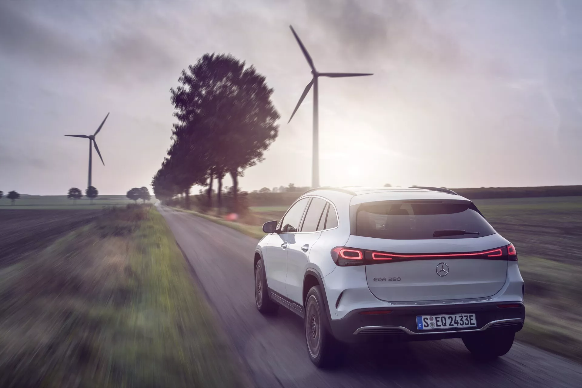 SUV elétrico Mercedes-Benz EQA 2021 em movimento, vista de traseira e lateral destacando design aerodinâmico e tecnologia moderna, com paisagem de turbinas eólicas de energia renovável ao fundo