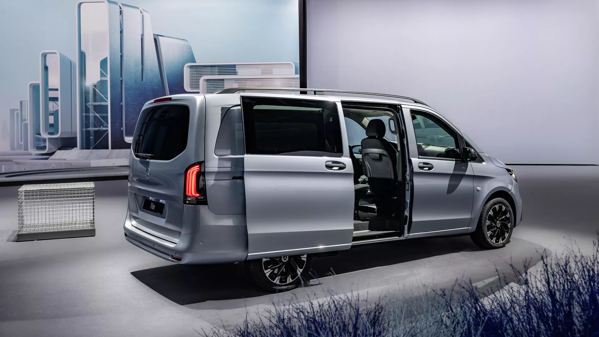 Minivan prata Mercedes-Benz Vito 2024 (447 facelift) com porta lateral deslizante aberta mostrando o design moderno e elegante de um ângulo traseiro de três quartos, destacando o perfil aerodinâmico do veículo, luzes traseiras LED, rodas de liga leve e um vislumbre do interior avançado com material de assento potencialmente premium e um painel moderno.