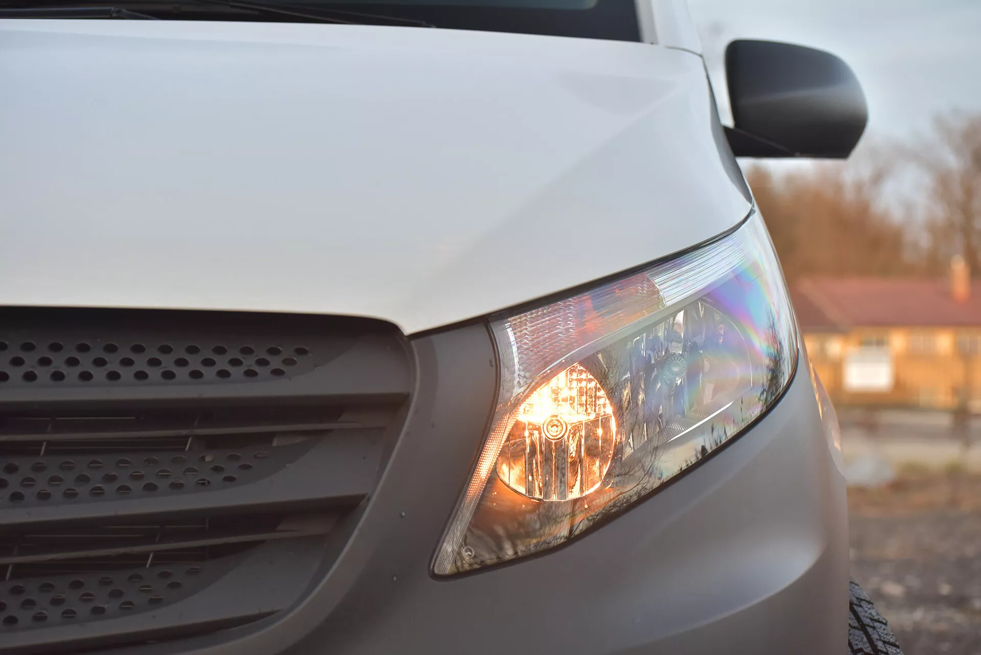 Vista lateral de perto de um Mercedes-Benz Vito Tourer branco de 2014 focando no farol iluminado, parte da grade e seção do capô, destacando seus faróis halógenos e design aerodinâmico sob iluminação suave.