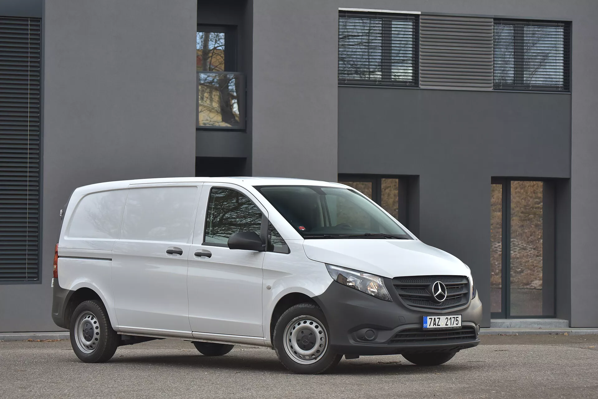 Furgão Mercedes-Benz Vito Tourer 2014 na cor branca estacionado no asfalto, exibindo perfil frontal e lateral com visão clara do emblema da grade e faróis modernos, fotografado em luz diurna nublada em frente a um edifício minimalista