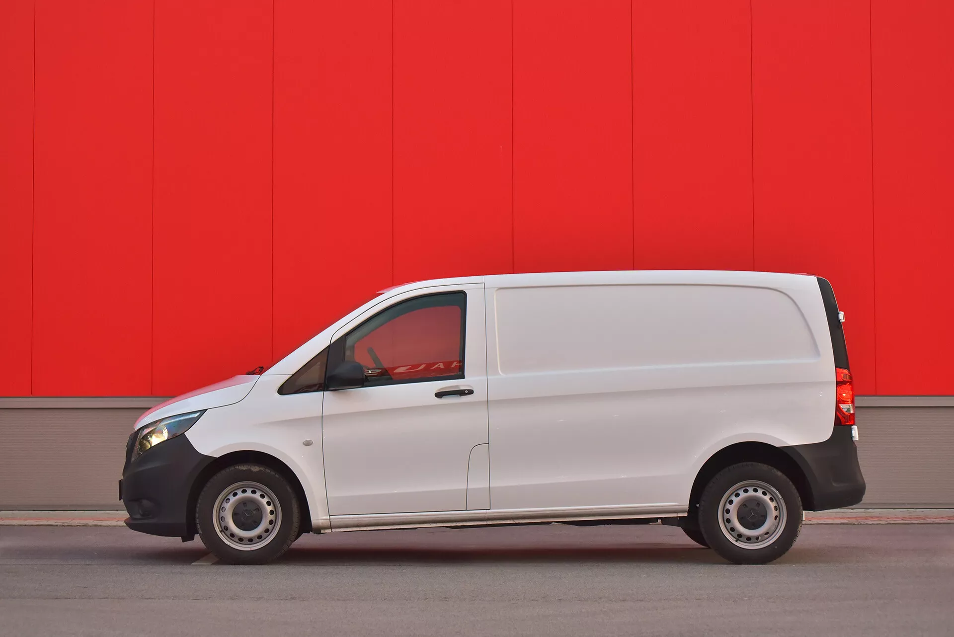Furgão comercial branco Mercedes-Benz Vito Tourer de 2014 estacionado paralelamente contra uma parede vermelha vibrante, mostrando perfil lateral com porta visível, janelas e rodas de aço capturadas de um ângulo baixo para enfatizar o design e as características do veículo