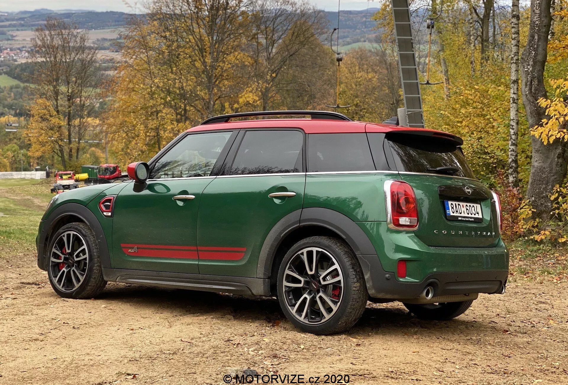 Vista traseira de três quartos do Mini Cooper Countryman verde 2020 com edição esportiva John Cooper Works, mostrando teto vermelho e tampas dos retrovisores, ponteiras de escape duplas, spoiler traseiro, rodas de liga leve de cinco raios com detalhes em preto e recursos tecnológicos avançados como sensores de estacionamento e antena tipo barbatana de tubarão, posicionado em uma superfície de terra em um ambiente rural de outono.