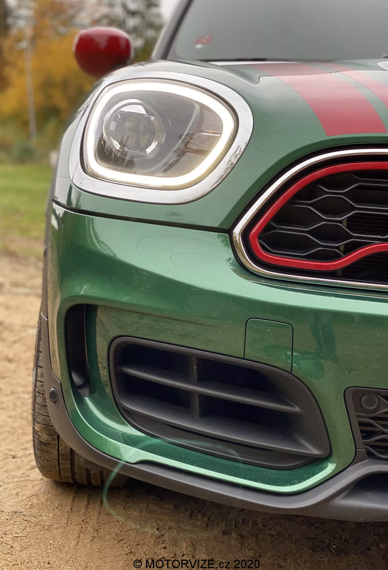 Imagem em close-up de um Mini Countryman John Cooper Works 2020 verde vibrante, focada no quarto frontal-esquerdo. Capturada de um ângulo baixo, a imagem destaca a grelha inferior com design de favo de mel preto, o para-choque dianteiro com características aerodinâmicas e o farol LED elegante. A grelha superior é hexagonal com acabamento preto brilhante e borda acentuada em vermelho. O carro possui faixas de corrida no capô e no teto, um espelho lateral vermelho visível no canto superior esquerdo, e está situado ao ar livre com luz natural, com árvores e folhagens desfocadas no plano de fundo.