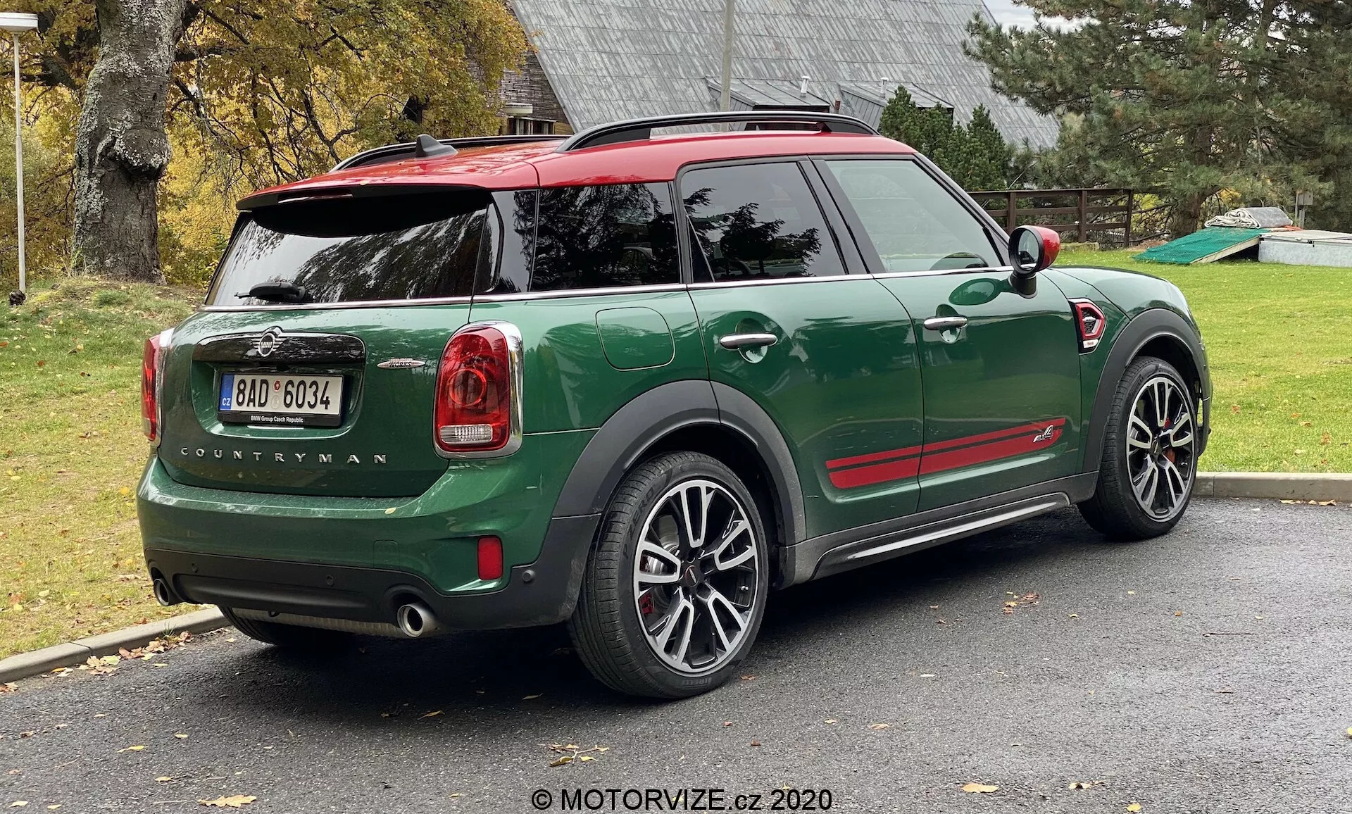 Vista traseira de três quartos de um MINI Countryman John Cooper Works 2020 verde estacionado em uma entrada residencial. A imagem mostra a metade traseira completa do lado direito do carro, incluindo a tampa traseira, roda traseira direita, parte do teto e as portas dos passageiros. O design esportivo e compacto do SUV apresenta acentos vermelhos contrastantes nas capas dos retrovisores laterais e no teto, arcos das rodas pretos e grandes rodas de liga leve multirraiadas. As luzes traseiras, o nome do modelo 'Countryman' e o logotipo MINI são claramente visíveis. O carro possui um teto vermelho com um spoiler e trilhos de teto, e o vidro traseiro inclui um desembaçador e um limpador.