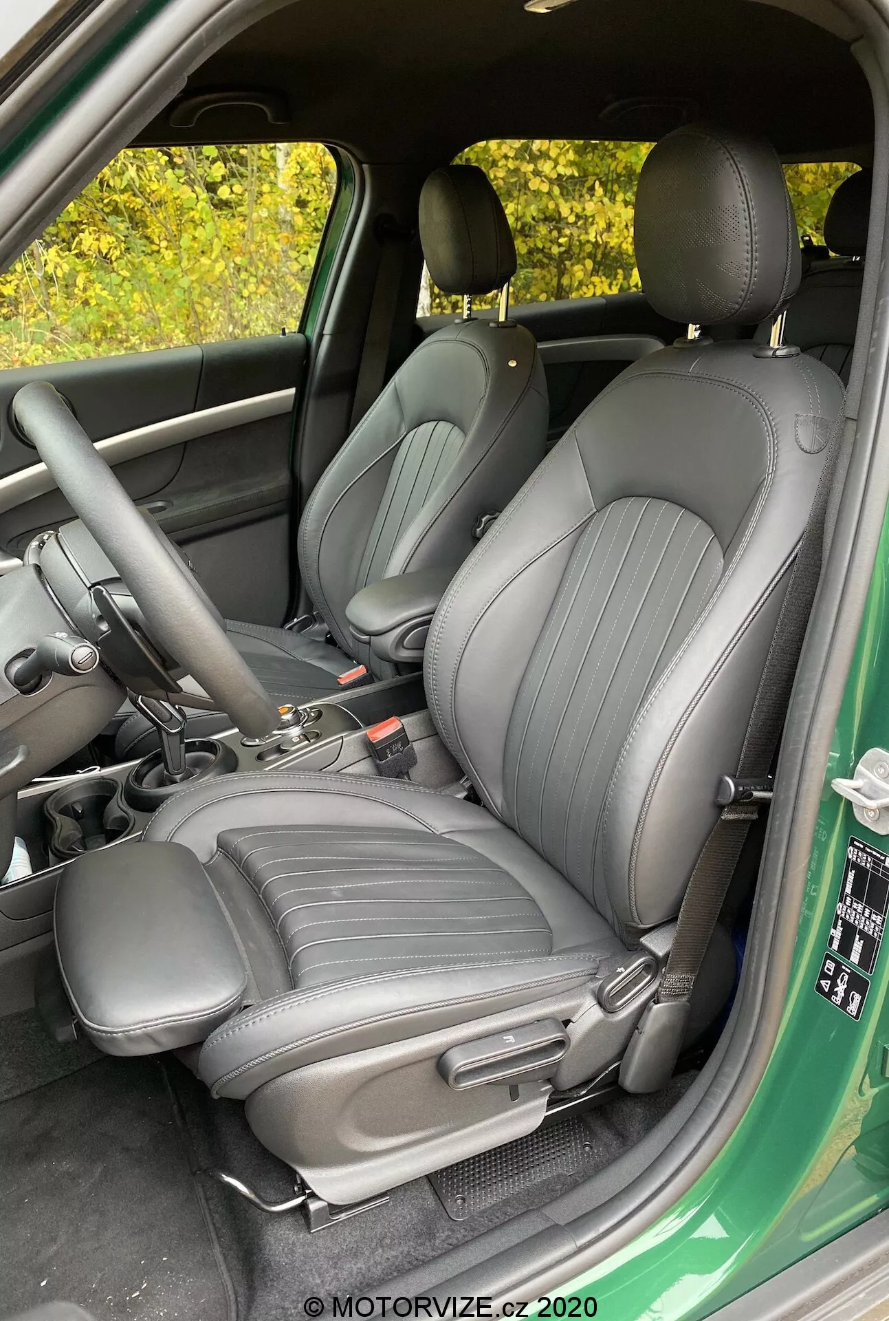 Imagem do interior do Mini Countryman John Cooper Works 2020, focada nos assentos dianteiros do condutor e do passageiro, estofados em material escuro de alta qualidade com linhas verticais, vista a partir da porta do condutor aberta, mostrando a consola central com alavanca de mudanças e travão de mão, vista parcial do painel da porta com controles para janelas e espelhos.