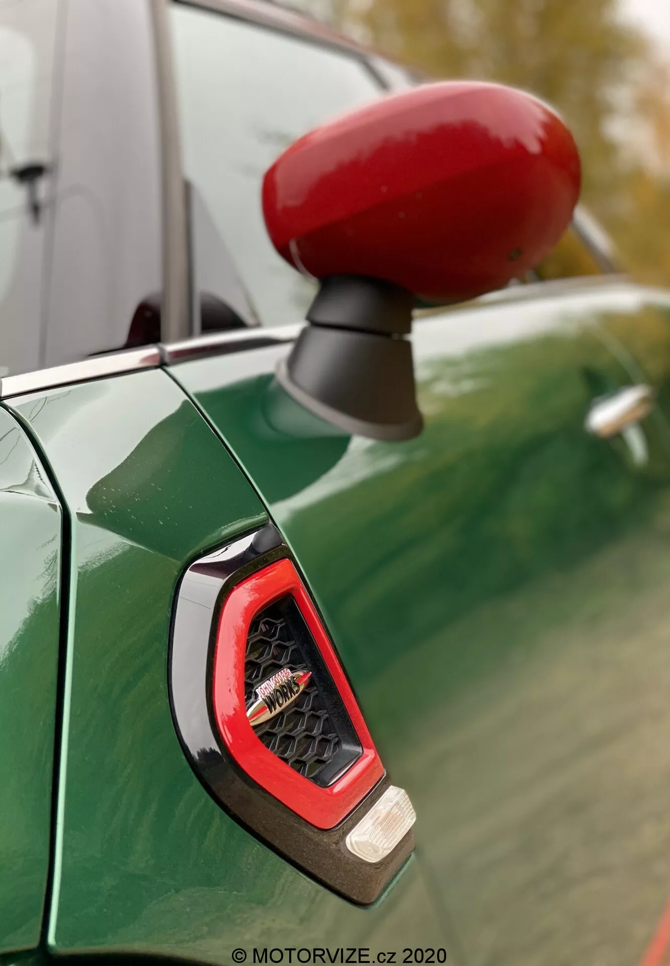 Close-up do espelho lateral de cor vermelha vibrante e do indicador lateral do lado do condutor de um Mini Countryman John Cooper Works 2020, visto contra um painel de carroçaria verde metálico profundo com reflexos de um céu nublado e árvores. O foco acentua o indicador angular com lente transparente, provavelmente exibindo tecnologia LED, rodeado por um acabamento esportivo e brilhante.