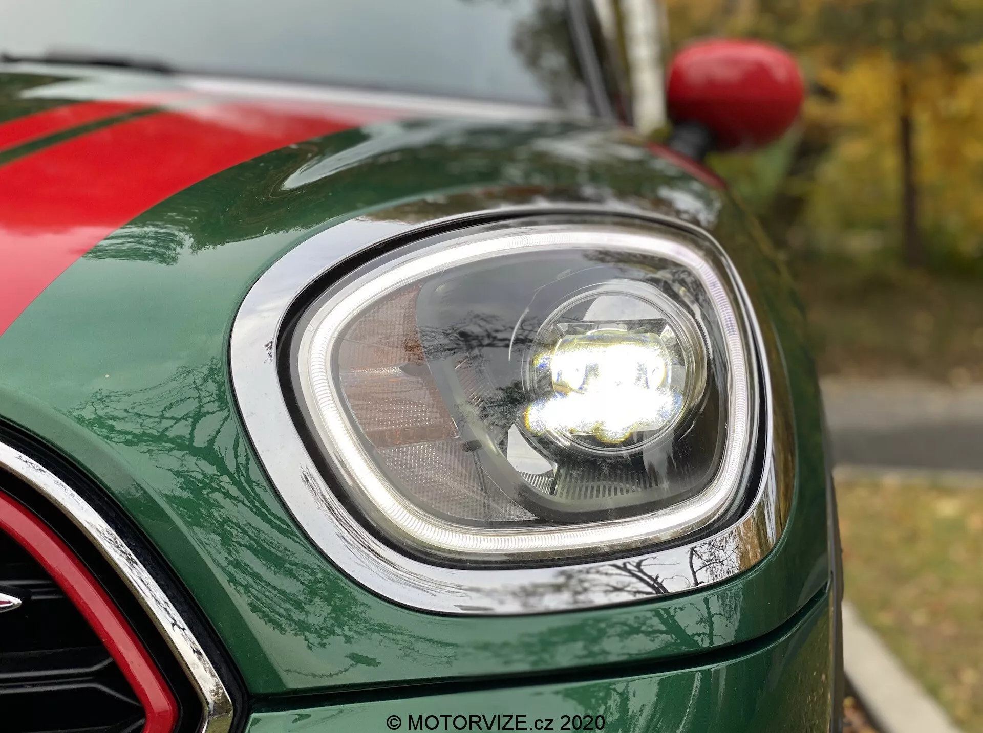 Vista aproximada da seção frontal esquerda de um Mini Countryman John Cooper Works 2020, focando no moderno farol redondo com tecnologia LED, tampa de lente transparente e farol de estilo projetor iluminado. A imagem mostra a curvatura do para-lama frontal, a borda do capô e parte da grade com acabamento cromado. O carro possui uma pintura verde metálica brilhante com flocos metálicos visíveis.