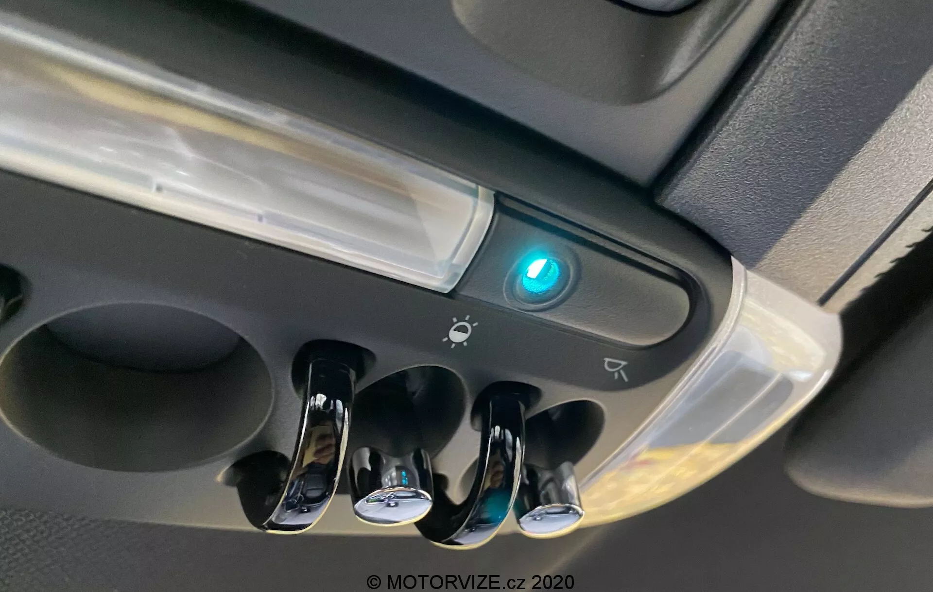 Vista aproximada da área do console superior do Mini Countryman 2020 John Cooper Works mostrando as luzes internas da cabine, três interruptores com acabamento cromado para luzes de leitura individuais, uma luz central da cabine, um botão iluminado em azul que pode indicar conectividade Bluetooth ou recurso de segurança, a borda de um possível compartimento de armazenamento, ícones sugerindo controles adicionais ou sensores e um design moderno com acabamentos brilhantes e foscos.