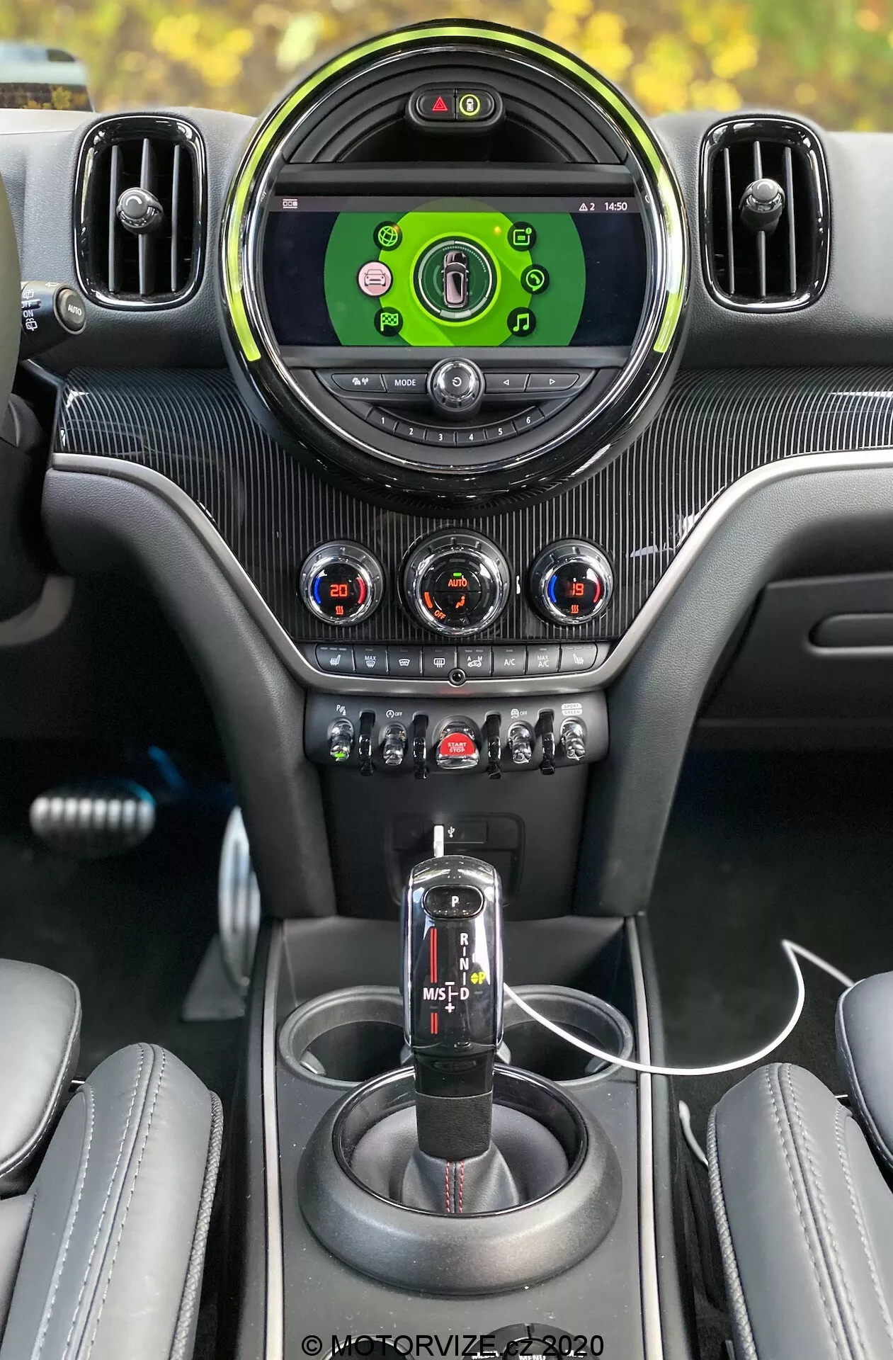 Vista_interna_de_um_Mini_Countryman_John_Cooper_Works_2020_da_perspectiva_do_motorista,_destacando_as_características_premium_como_a_grande_tela_circular_do_sistema_de_informação_e_entretenimento_com_interface_verde,_cercada_por_saídas_de_ar_ajustáveis_com_borda_prateada;_três_botões_de_controle_climático_com_indicadores_vermelhos_e_azuis_no_painel_texturado_de_fibra_de_carbono;_uma_fila_de_botões_e_interruptores_para_várias_funções_do_carro,_incluindo_modo_esporte/tração;_alavanca_de_câmbio_automática_preta_com_opção_de_modo_manual;_console_central_coeso_com_acabamento_em_fibra_de_carbono;_dois_suportes_para_bebidas;_um_cabo_USB_conectado_para_carregamento;_e_bancos_de_couro_parcialmente_visíveis_com_costuras,_tudo_combinado_para_criar_um_interior_automotivo_premium_centrado_na_tecnologia.