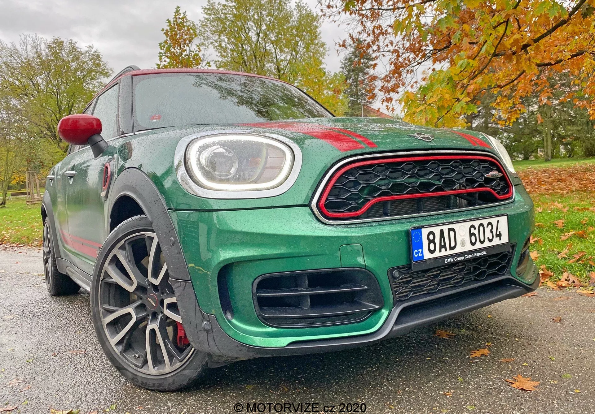 A imagem apresenta o Mini Countryman 2020 edição John Cooper Works a partir de um ângulo frontal baixo, destacando seu design esportivo e moderno. Os elementos visíveis incluem a grelha frontal em padrão de favo de mel com detalhes em vermelho, grandes entradas de ar abaixo da grelha para melhorar o fluxo de ar, uma placa de licença central e luzes diurnas LED circulares embutidas nos faróis principais. O capô apresenta uma ligeira saliência com dobras aerodinâmicas, enquanto as linhas esculpidas no perfil lateral melhoram seu aspecto dinâmico. As rodas de liga leve multirraiadas sugerem uma construção leve para melhor manuseio e aceleração, e revelam discos de freio de alto desempenho. A carroceria é pintada em um rico verde metálico, acentuada por uma capa de espelho lateral vermelho e detalhes vermelhos na grelha, contra um fundo outonal de folhas. Esta perspectiva enfatiza as características tecnológicas e de design avançadas do carro, fazendo com que se destaque nas categorias de esportivos e carros de luxo.