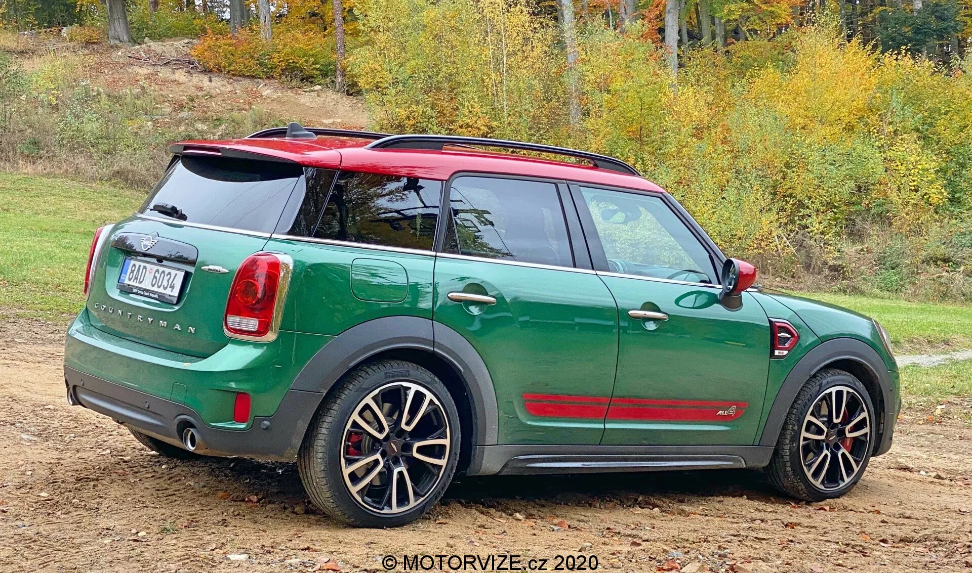 Vista traseira três quartos de um Mini Countryman John Cooper Works 2020 estacionado em uma superfície não pavimentada com manchas de grama. O compacto esportivo possui um esquema de cores duplo, com carroceria em verde escuro e teto e espelhos em vermelho contrastante. O design traseiro exibe uma porta traseira, limpador de para-brisa traseiro, emblema proeminente e lanternas traseiras dispostas verticalmente. O para-choque traseiro inclui sensores de estacionamento. Ele também possui rodas de liga leve esportivas com design de múltiplos raios que mostram pinças de freio, sugerindo uma versão de alto desempenho. O fundo natural com árvores outonais sugere a estação.