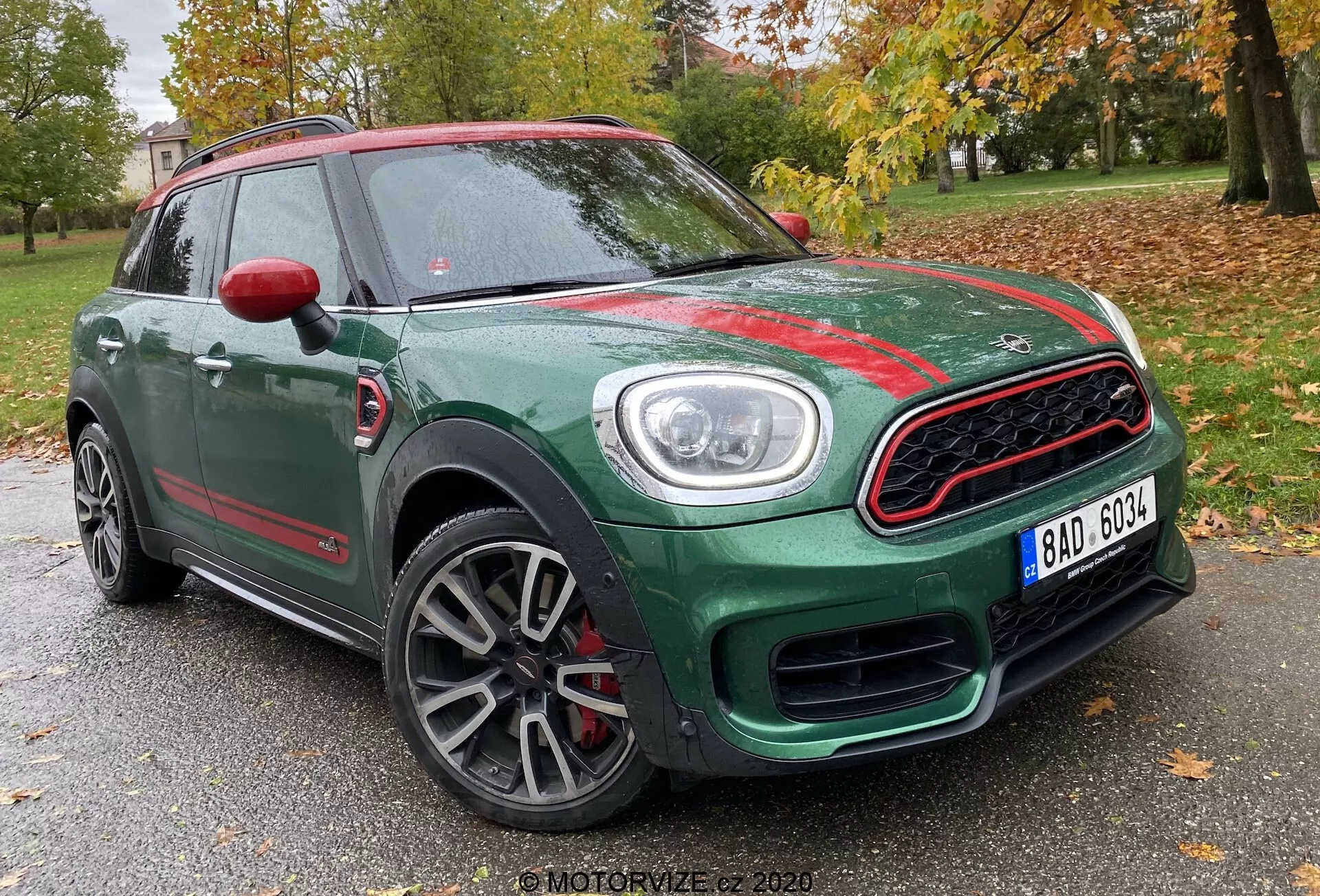 Vista de três quartos frontal do Mini Countryman John Cooper Works 2020, mostrando sua carroceria compacta de SUV verde com detalhes contrastantes em vermelho. Capturada de um ângulo baixo, a imagem destaca a grade frontal esportiva com contorno cromado e design de colméia, os faróis circulares proeminentes com luzes diurnas de LED, as rodas de liga leve de múltiplos raios com acabamento cinza e as capas dos espelhos laterais vermelhas. A foto foi tirada com um fundo de árvores e uma estrutura tênue, sugerindo uma localização em um parque ou área residencial.