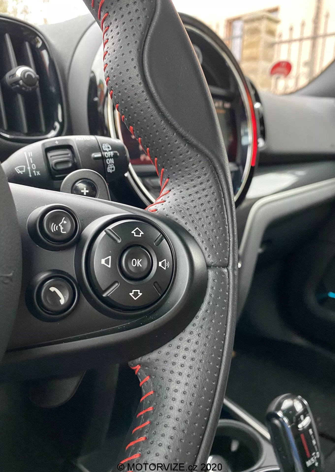 Vista interior de um Mini Countryman John Cooper Works 2020 focada no volante revestido em couro com costura vermelha, botões de controle para áudio e chamadas, e painel de instrumentos parcialmente visível com acentos vermelhos indicando um modelo esportivo. Mostra a área de controle do motorista, a alavanca de câmbio automática e os controles dos vidros elétricos, destacando conveniência e funcionalidades avançadas.