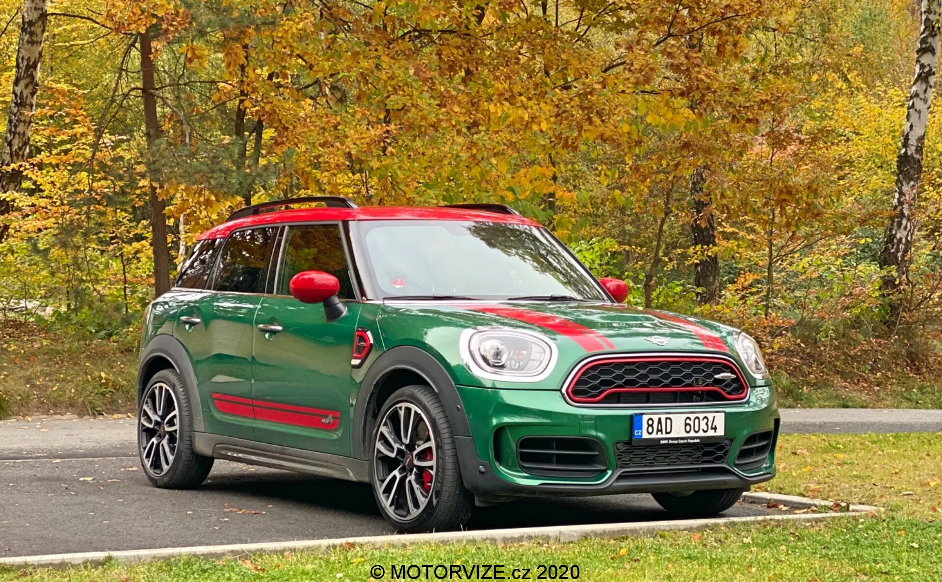Um Mini Countryman John Cooper Works 2020 verde capturado desde uma vista frontal de três quartos. A fotografia mostra tanto a frente quanto o lado do carro, destacando suas características de design. A parte frontal tem uma grade distinta com uma placa de licença abaixo e faróis redondos clássicos. Faixas esportivas correm ao longo do capô, e o perfil lateral mostra rodas de liga, espelhos laterais e a linha do teto. Espelhos laterais vermelhos e um teto combinando contribuem para um esquema de cores de dois tons. Abaixo das portas, os estribos laterais coordenados sugerem um nível de acabamento ou pacote esportivo atualizado. O veículo possui um teto solar panorâmico e está estacionado em uma superfície de asfalto com um fundo natural de outono. A foto nítida e brilhante foi tirada com luz natural.