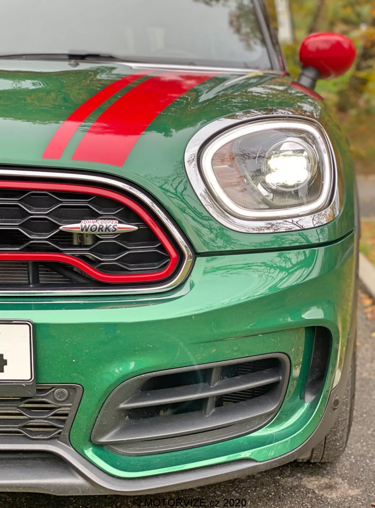Vista aproximada do quarto dianteiro esquerdo de um Mini Countryman John Cooper Works 2020. A imagem, tirada ligeiramente abaixo do nível dos faróis, destaca o capô metálico verde com uma faixa de corrida vermelha, o farol esquerdo circular cercado por cromo e uma parte da grade em favo de mel com um contorno preto brilhante. É visível a insígnia 'John Cooper Works', o indicador esquerdo na moldura da roda e as grandes entradas de ar no para-choque dianteiro, projetadas para o resfriamento do motor e desempenho aerodinâmico. O carro parece limpo e bem conservado, destacando sua tecnologia moderna de faróis de projeção ou LED e design esportivo orientado ao desempenho.
