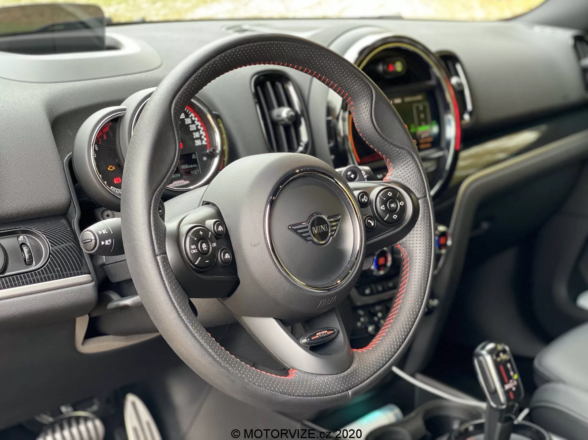 Vista interior do Mini Countryman John Cooper Works 2020 da perspectiva do condutor, com volante de fundo plano revestido em couro com costuras visíveis e controles multifuncionais, palhetas de mudança atrás do volante, dois mostradores circulares de instrumentos flanqueando um display digital central, e um painel com vários botões e interruptores no console central.