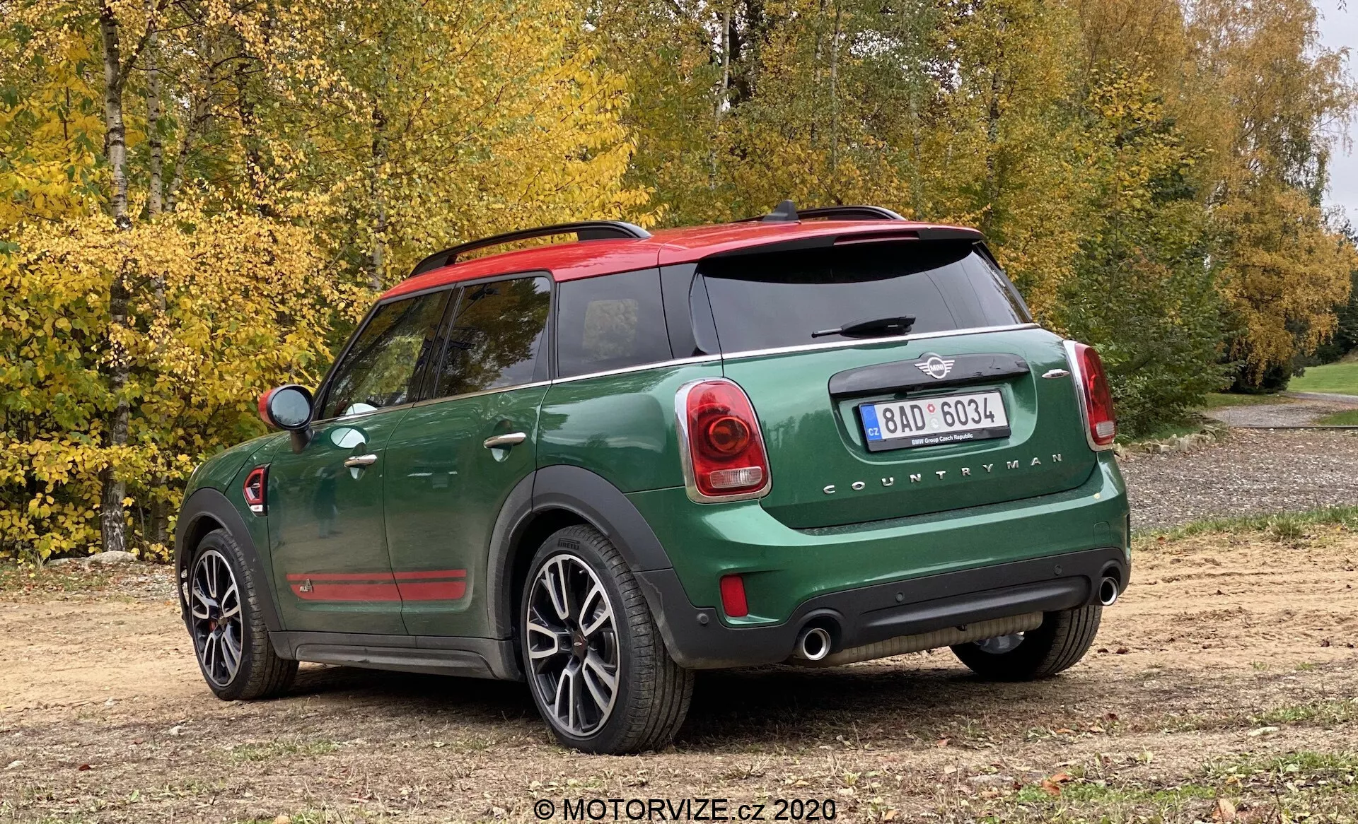 Vista traseira de três quartos de um Mini Cooper Countryman John Cooper Works 2020 com pintura de dois tons verde escuro e preto, capas dos espelhos vermelhas, emblema Mini visível e letras 'Countryman', luzes traseiras de design único, para-choque traseiro proeminente com sensores de estacionamento, janela traseira com desembaçador, tubos de escape duplos, limpador traseiro, rodas de liga leve de cinco raios, estacionado em uma estrada de terra com árvores e folhas de outono ao fundo, capturado em um ângulo ligeiramente alto.