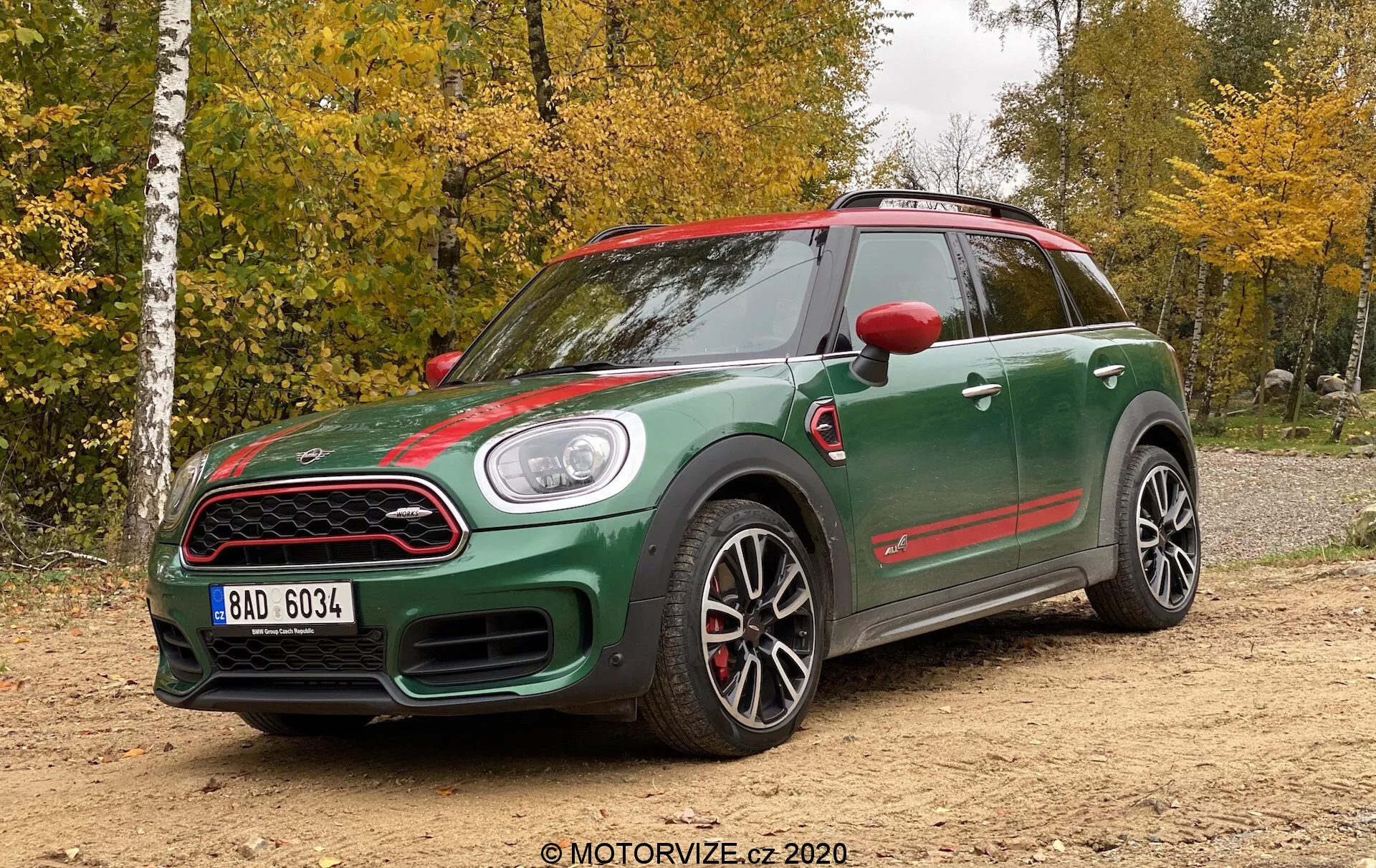 Vista angular de três quartos da frente de um Mini Countryman John Cooper Works 2020, exibindo seu design esportivo com carroceria bicolor verde escuro e detalhes vermelhos nos retrovisores, grade frontal e saias laterais. Os recursos incluem um scoop distintivo no capô, faróis redondos, grade frontal em favo de mel, grandes rodas esportivas de liga leve com design multirraios, pinças de freio vermelhas, saias laterais pronunciadas e uma postura agressiva. O carro está posicionado em uma superfície não pavimentada com uma vegetação de floresta ao fundo, sob luz natural em um dia nublado.
