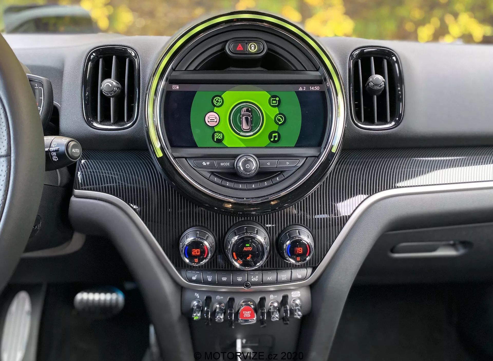 Painel interior do Mini Countryman John Cooper Works 2020 visto da perspectiva do motorista. Console central com grande tela de infotainment circular mostrando ícones coloridos para mídia, conectividade telefônica, navegação e configurações do veículo. Enquadrado por uma moldura circular elegante e flanqueado por saídas de ar adicionais. Abaixo da tela, há mostradores analógicos com detalhes cromados para controle climático e interruptores para funções como aquecimento dos assentos, descongelamento, recirculação de ar e modos de condução. Textura semelhante à fibra de carbono e detalhes metálicos fornecem um design interior moderno e luxuoso com painel de instrumentos digital visível indicando que o veículo está ligado.