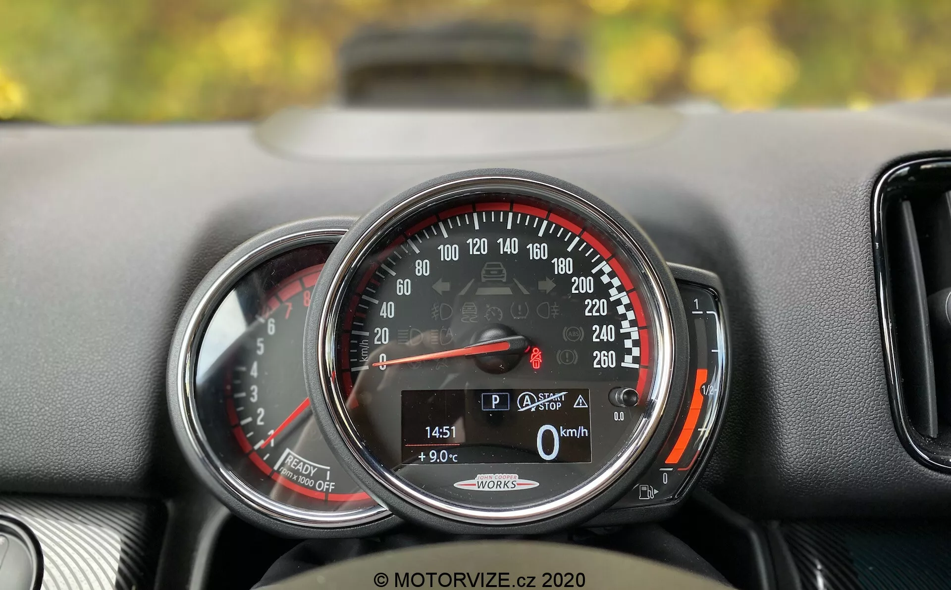 Close-up detalhado do painel de instrumentos do Mini Countryman John Cooper Works 2020. O foco está em um velocímetro circular com uma escala de 0 a 260 km/h, com o ponteiro indicando 0 km/h, sugerindo que o carro está parado. Um visor digital na parte inferior mostra o carro no modo 'P', uma temperatura externa de 9,0°C, uma leitura de quilometragem de 1451 km e um ícone de porta-malas aberto. As palavras 'Auto Start/Stop' sugerem um sistema de economia de combustível. O velocímetro é emoldurado por um anel de cromo e possui uma zona vermelha começando pouco depois de 220 km/h. À esquerda, há uma visão parcial de outro medidor, provavelmente um tacômetro. O painel tem uma superfície texturizada e uma pequena ventilação de ar é parcialmente visível à direita do velocímetro.