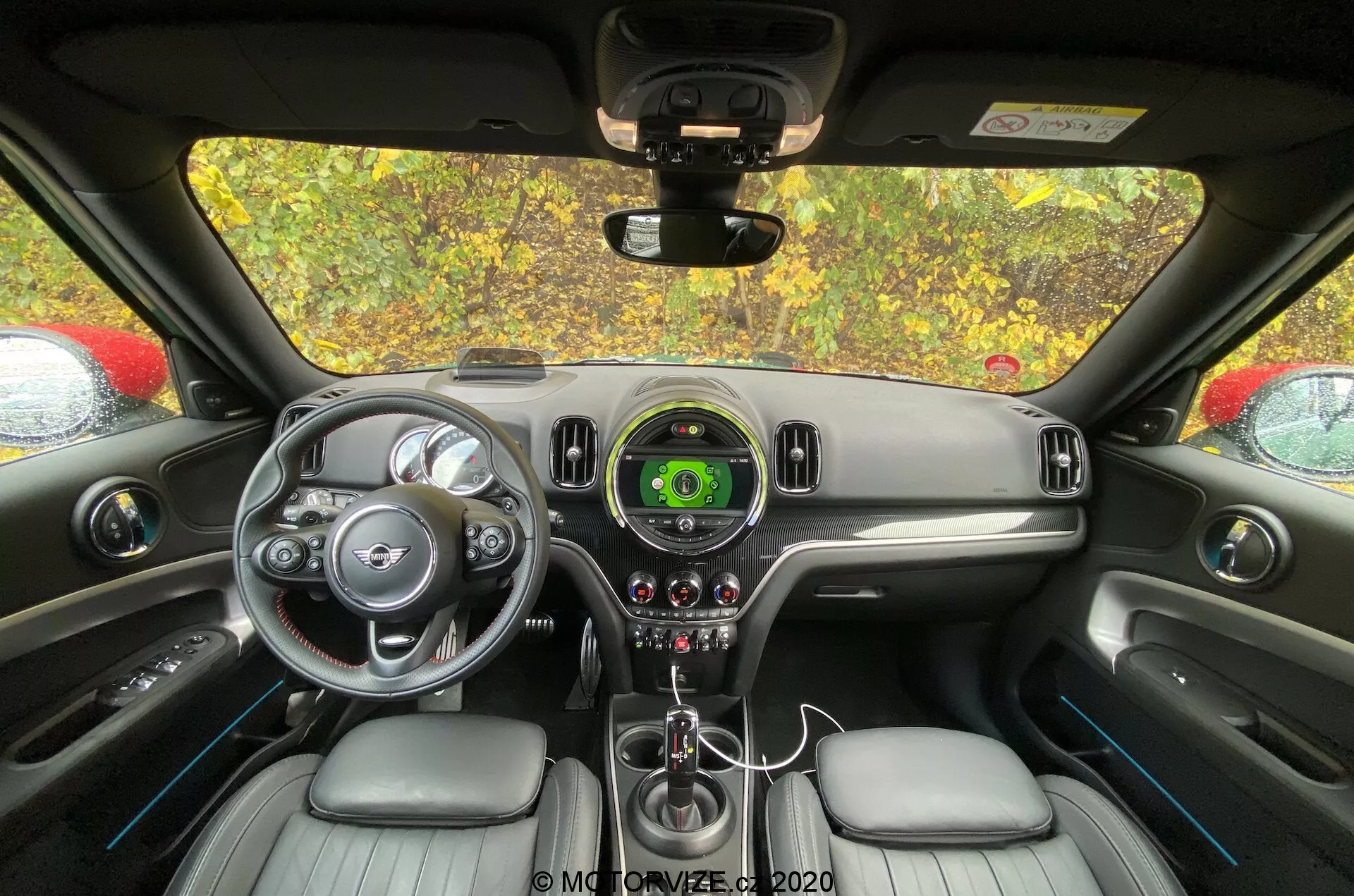 Interior do Mini Countryman 2020 John Cooper Works a partir de uma perspetiva do banco traseiro, focado no painel de instrumentos e área de controle do condutor. O volante com acentos prateados e logótipo da marca, o velocímetro central com ecrã digital, design elegante do painel de instrumentos, alavanca de câmbio, controles de clima, assentos estofados em couro e espelho retrovisor são visíveis.