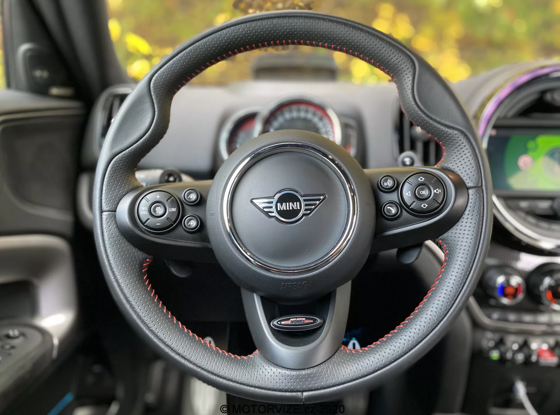Vista interior do Mini Countryman John Cooper Works 2020 a partir do banco do motorista, com foco no volante com o logotipo Mini no centro. O volante de três raios, revestido em couro com costuras vermelhas, possui controles multifuncionais para áudio, telefone, controle por voz e controle de cruzeiro. As aletas de mudança são visíveis, indicando uma transmissão automática com seleção manual de marchas. No fundo, pode-se ver o painel com elementos de design circulares, um display central mostrando um mapa, saídas de ar-condicionado e detalhes coloridos.