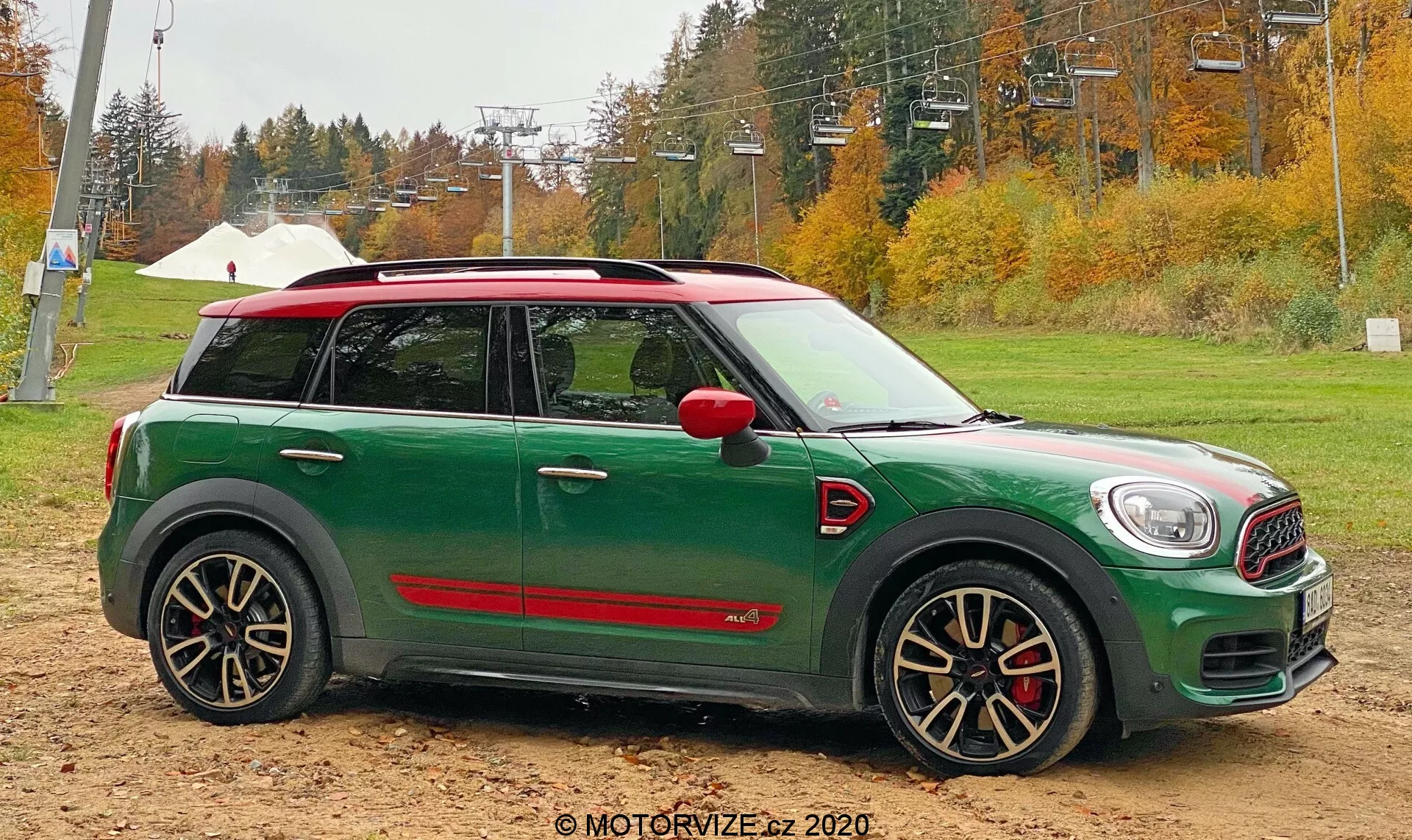 Perfil lateral de um Mini Cooper Countryman 2020 John Cooper Works verde, posicionado em solo arenoso com árvores de outono e infraestrutura de teleférico ao fundo. O carro possui teto branco, capas dos retrovisores vermelhas, rodas pretas e prateadas agressivas com vários raios e pinças de freio vermelhas, e saias laterais esportivas vermelhas.