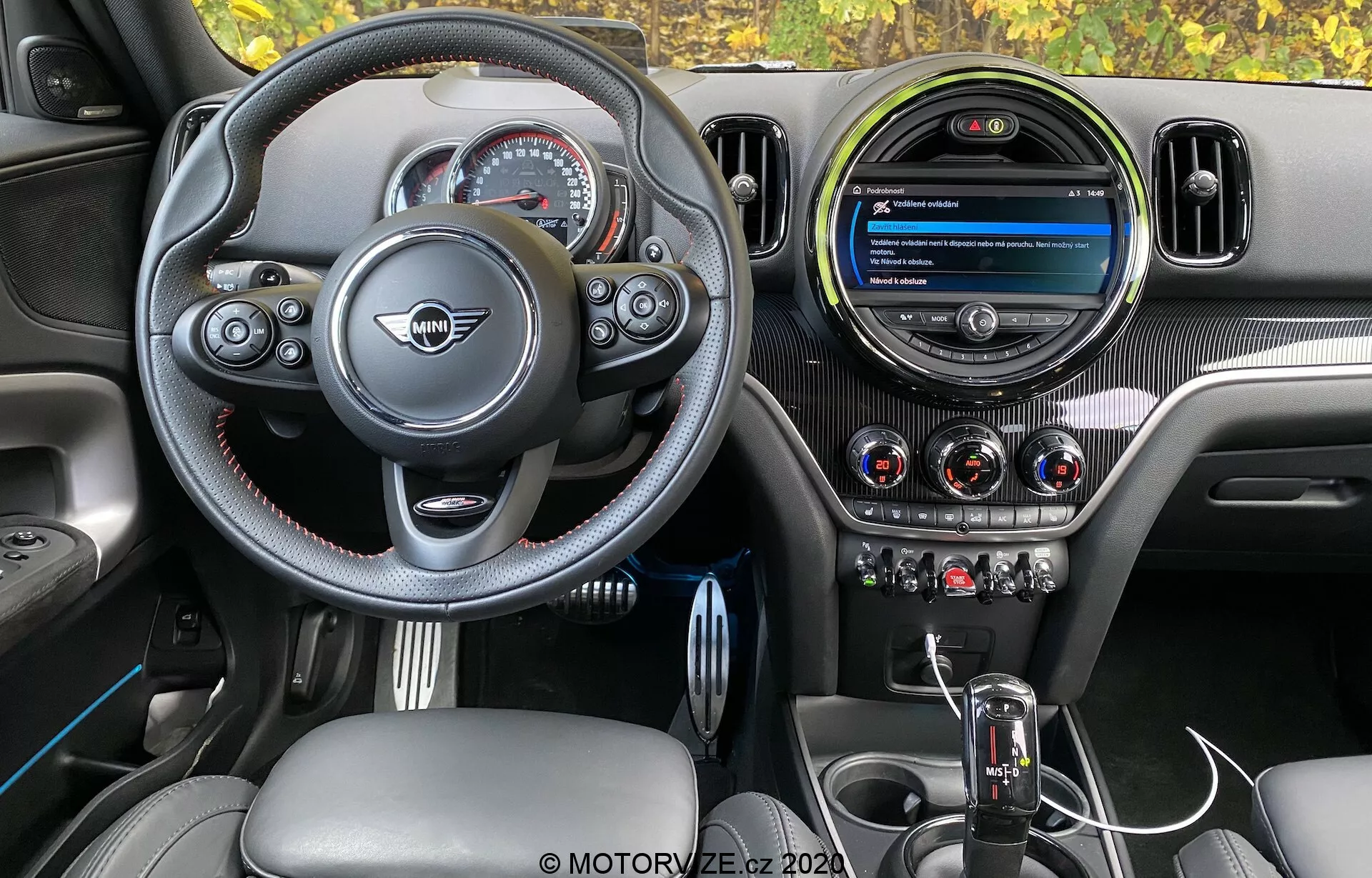 Vista interior do Mini Countryman John Cooper Works 2020, destacando o cockpit do motorista com volante esportivo de fundo plano, botões multifuncionais, costuras de contraste e emblema central do fabricante. O painel de instrumentos inclui uma tela circular de infotainment moderna, três saídas de ar metálicas e botões de controle de clima integrados. A consola central apresenta uma alavanca de câmbio automática com capa de couro e um botão de controle infotainment, todos complementados por texturas de fibra de carbono, couro e acabamentos metálicos para um design premium e orientado para o desempenho.