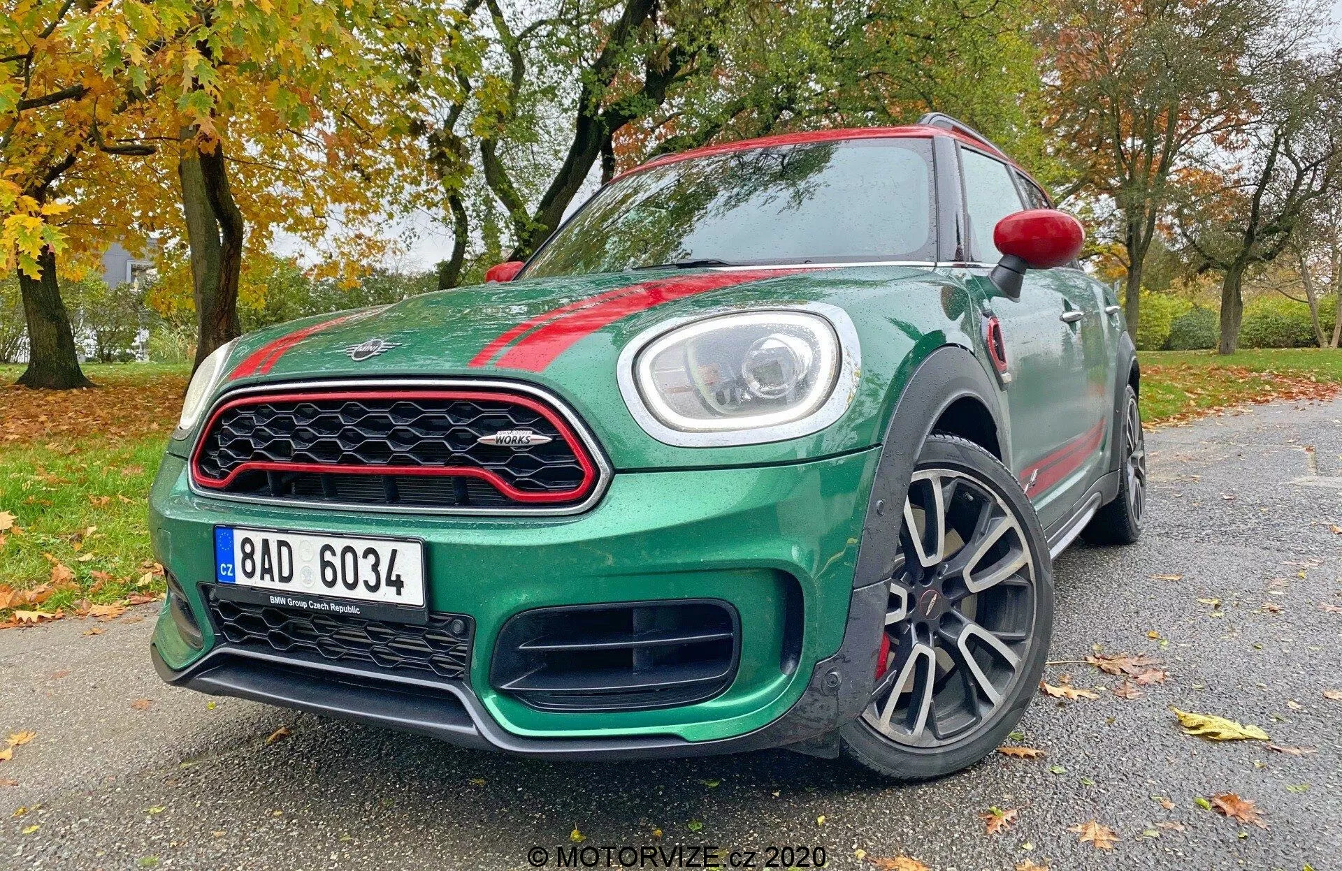 A imagem mostra um hatchback Mini Countryman John Cooper Works 2020, capturado de um ângulo baixo de três quartos frontal. São visíveis elementos-chave de design e tecnologia, incluindo faróis de LED, uma grade frontal distinta, um para-choque estilizado, rodas de liga leve, espelhos laterais parcialmente visíveis com indicadores de seta integrados, linhas aerodinâmicas do corpo, emblemas e o acabamento de pintura brilhante do carro. Esta perspectiva enfatiza a postura agressiva e o design dinâmico do veículo.