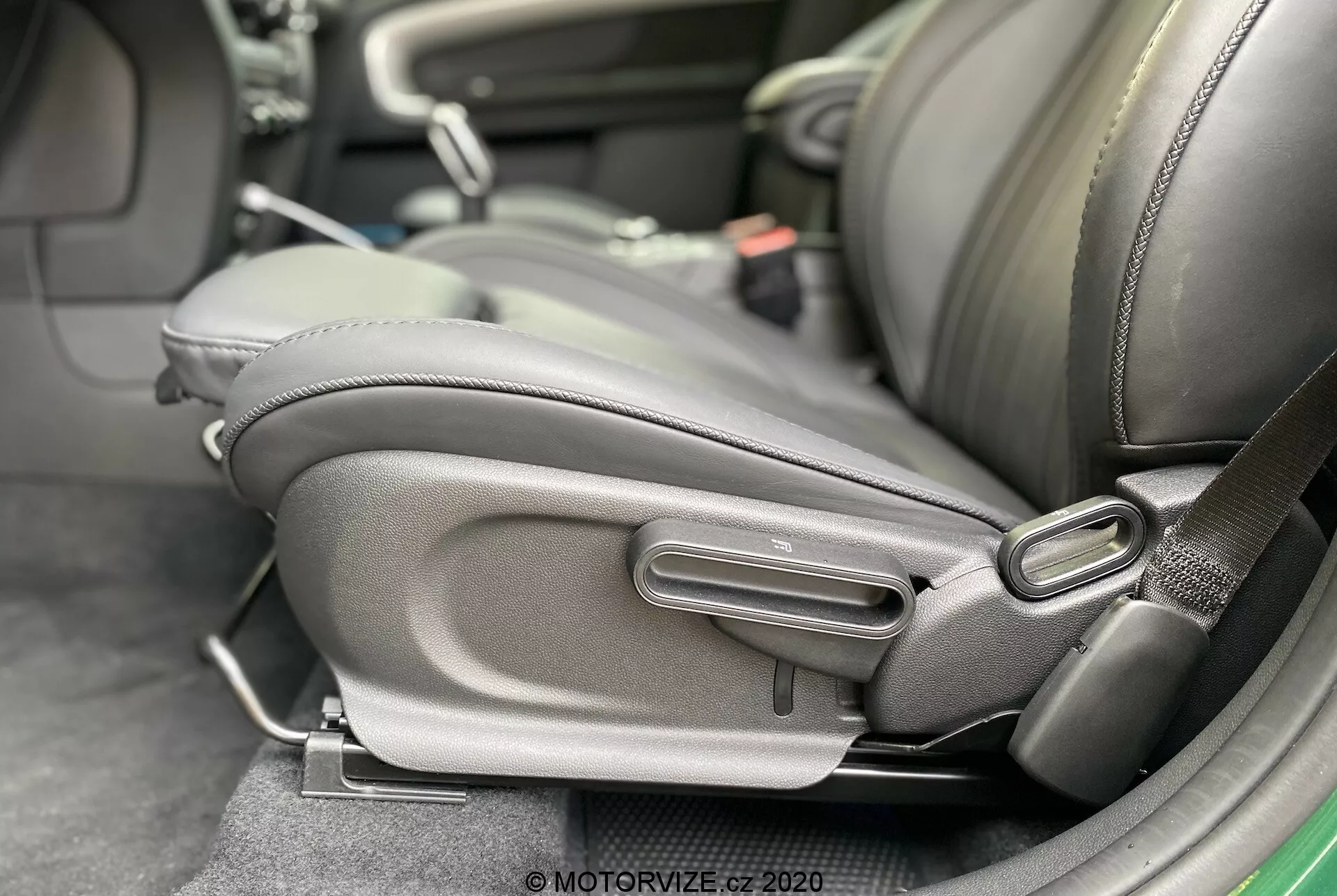 Vista de perto do interior do Mini Countryman John Cooper Works 2020, destacando a parte lateral inferior do banco do motorista com mecanismos de ajuste detalhados. O banco é estofado em couro escuro ou material semelhante a couro com um reforço lateral proeminente, focando-se no conforto e na qualidade. Controles manuais de ajuste são visíveis, incluindo uma alça de barra e um mostrador redondo, sugerindo um design ergonômico e ajustabilidade pelo usuário. Um receptáculo do cinto de segurança está presente, indicando atenção à segurança do passageiro.