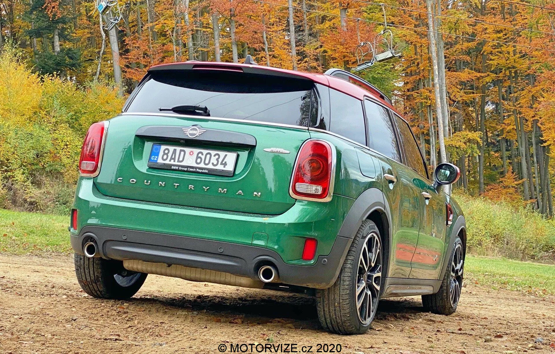 Imagem de um Mini Countryman 2020 modelo John Cooper Works verde estacionado ao ar livre em um cenário de outono, tirada de um ângulo traseiro de três quartos. O lado traseiro direito do carro está voltado para a câmera e mostra as luzes traseiras, o para-brisa traseiro, o emblema 'COUNTRYMAN', o sistema de escape duplo, as rodas de cinco raios, o teto preto contrastante e os espelhos laterais. A imagem também inclui uma placa traseira, limpador traseiro e antena para conectividade.