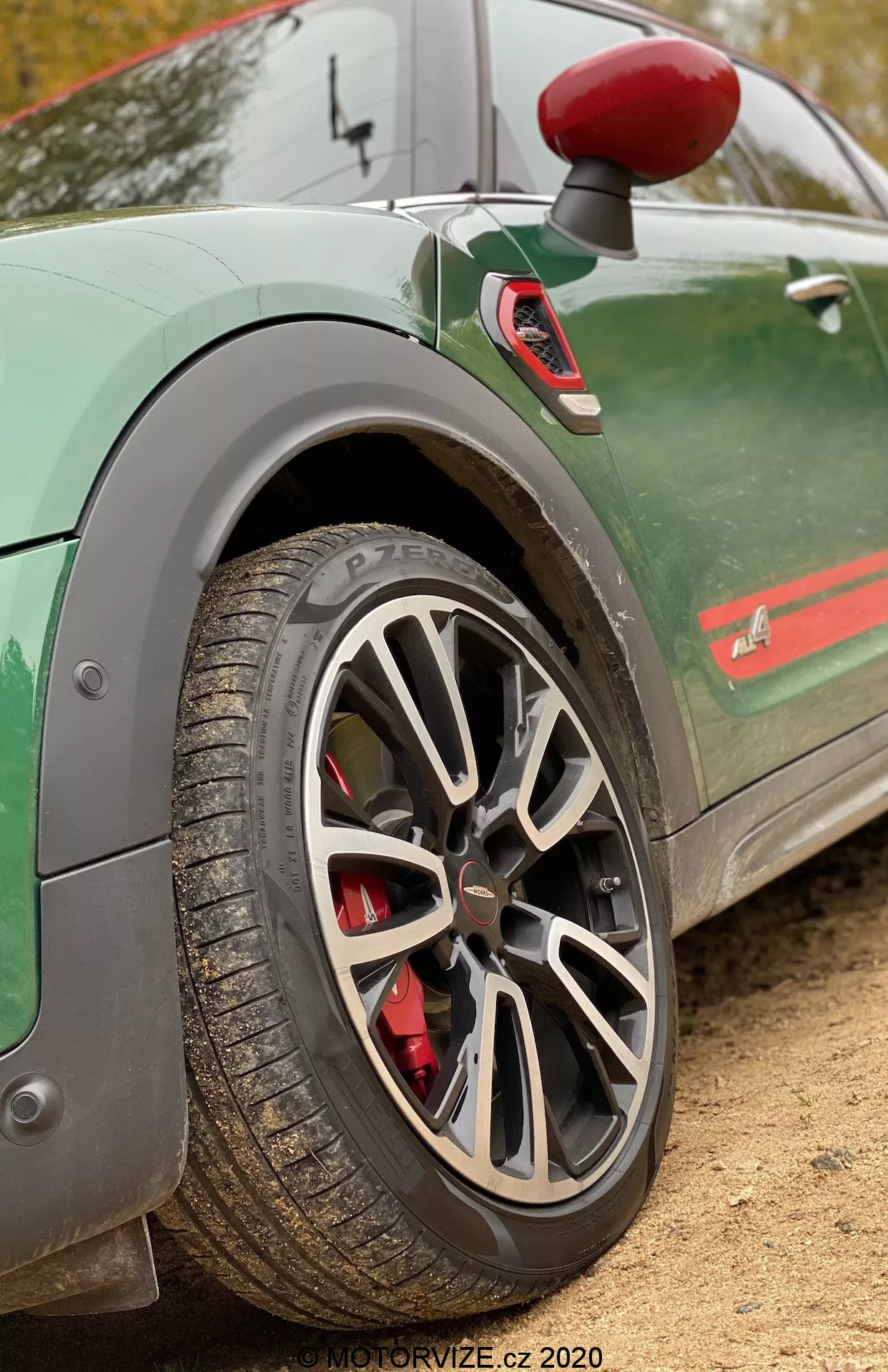 Imagem em close-up da roda traseira e parte do para-lama traseiro de um Mini Countryman John Cooper Works 2020. A foto é tirada de um ângulo lateral, ligeiramente inclinado e baixo, para enfatizar a roda de liga leve de múltiplos raios com acabamento cinza ou prata. A pinça de freio vermelha sugere um sistema de freio de alto desempenho. O pneu tem sulcos visíveis, indicando sua adequação para diversas condições de estrada. O carro verde apresenta uma faixa estilizada ao longo da parte inferior das portas e um emblema parcialmente visível que adiciona uma estética esportiva. Um espelho retrovisor lateral com capa vermelha e um para-barro também são visíveis, aumentando a aparência dinâmica do veículo. O fundo desfocado destaca esses detalhes, ideal para realçar elementos de desempenho automotivo.