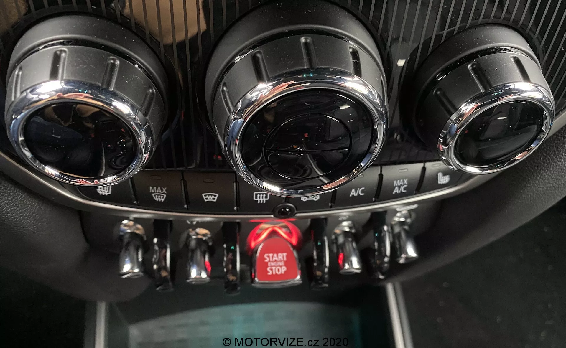 Esta imagem captura uma vista de close-up do console central do painel de instrumentos em um Mini Countryman John Cooper Works 2020, destacando o sistema de controle do clima e o botão de início/parada do motor. O console exibe duas saídas de ar circulares com acabamentos metálicos no topo, um painel de controle do clima detalhado com três botões metálicos para temperatura, velocidade do ventilador e direção do fluxo de ar, e vários botões para funções como degelo e recirculação de ar. Abaixo dos controles há um botão de início/parada do motor vermelho proeminente. As texturas do painel incluem um design esportivo semelhante a fibra de carbono ao redor das saídas de ar e plástico preto padrão para o painel inferior. O ângulo da foto, de frente e ligeiramente acima, enfatiza o design de alto padrão e os recursos avançados.