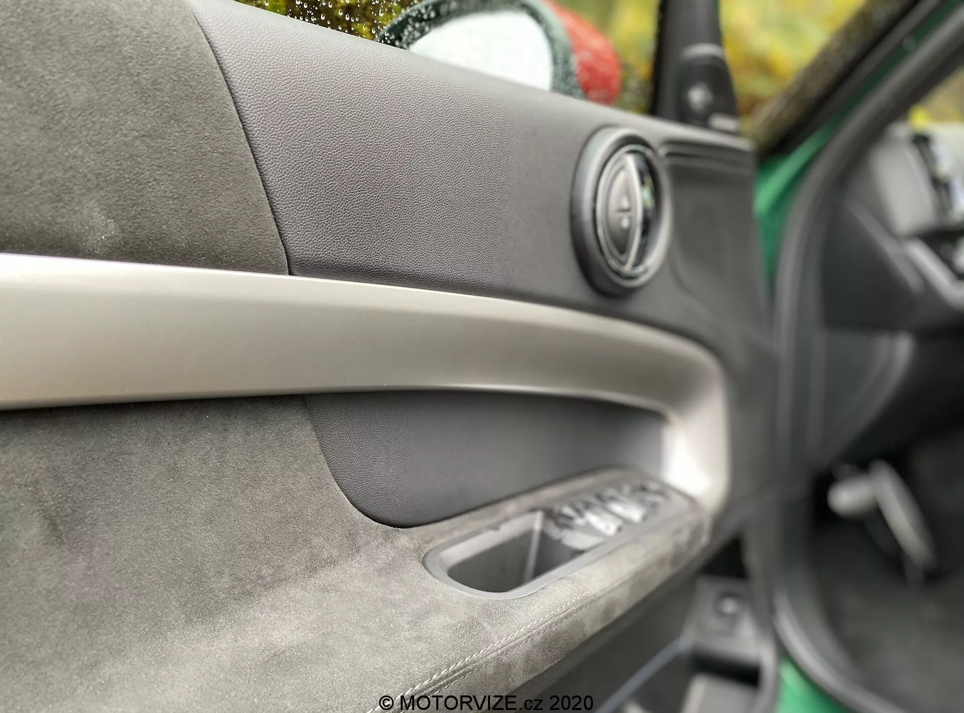 Imagem detalhada do interior da porta de um carro Mini Countryman John Cooper Works 2020. Foco no alto-falante premium com anel metálico, faixa decorativa diagonal, material macio ao toque com costuras visíveis, maçaneta interna e controles dos vidros elétricos. Profundidade de campo mínima destacando a arte e o conforto interno.